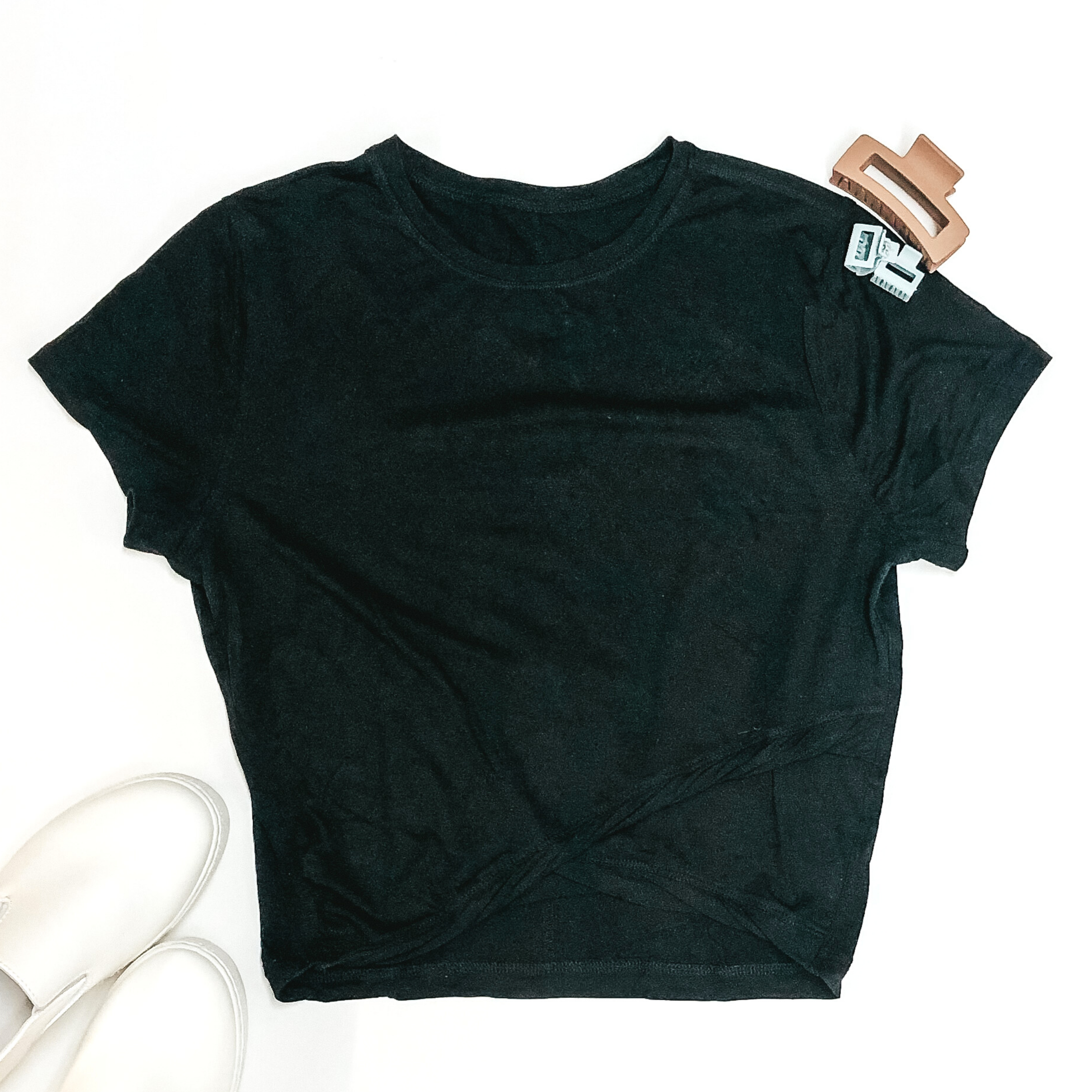 Short sleeve, cropped black tee. This tee is cropped, has a criss cross hemline, nad a scoop neckline. This tee is pictured on a white background with white shoes and hair clips in the background.