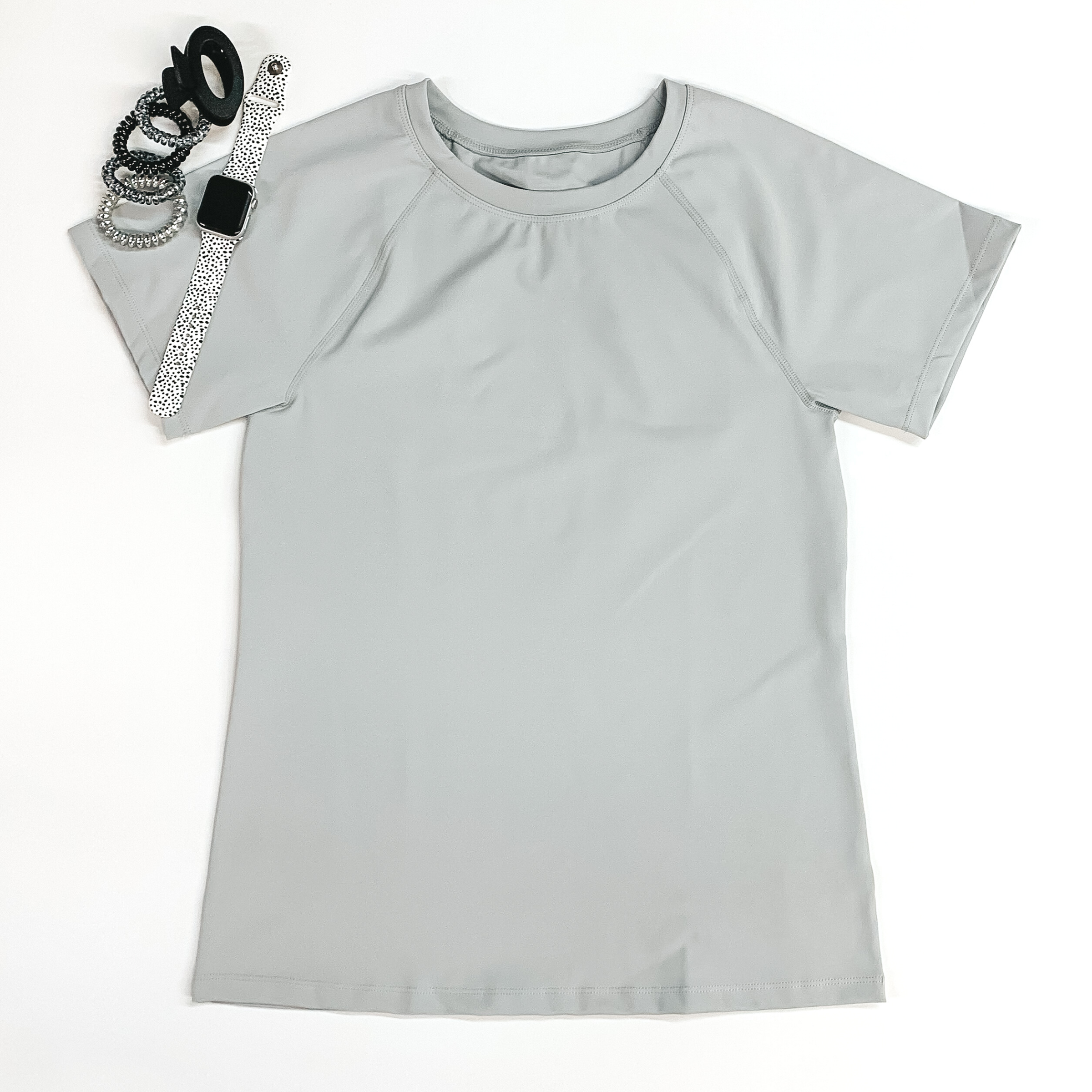 Light grey shorts sleeve top pictured on a white background with hair ties, a black clip, and a smart watch at the top left corner. 