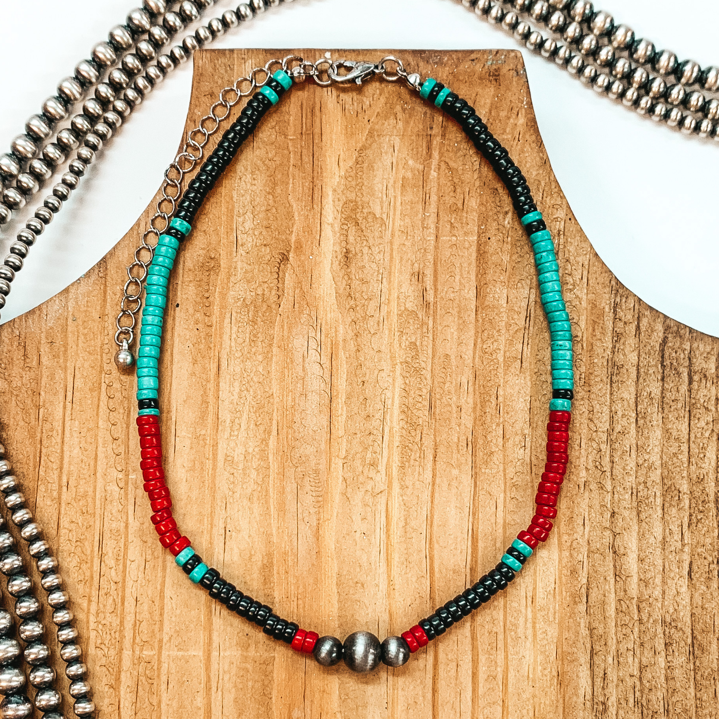 This necklace has segments of beads in the colors black, red, and turquoise with black beaded spacers. This necklace also includes three silver beads at the center. this necklace is pictured on a brown necklace holder on a white background with silver beads.