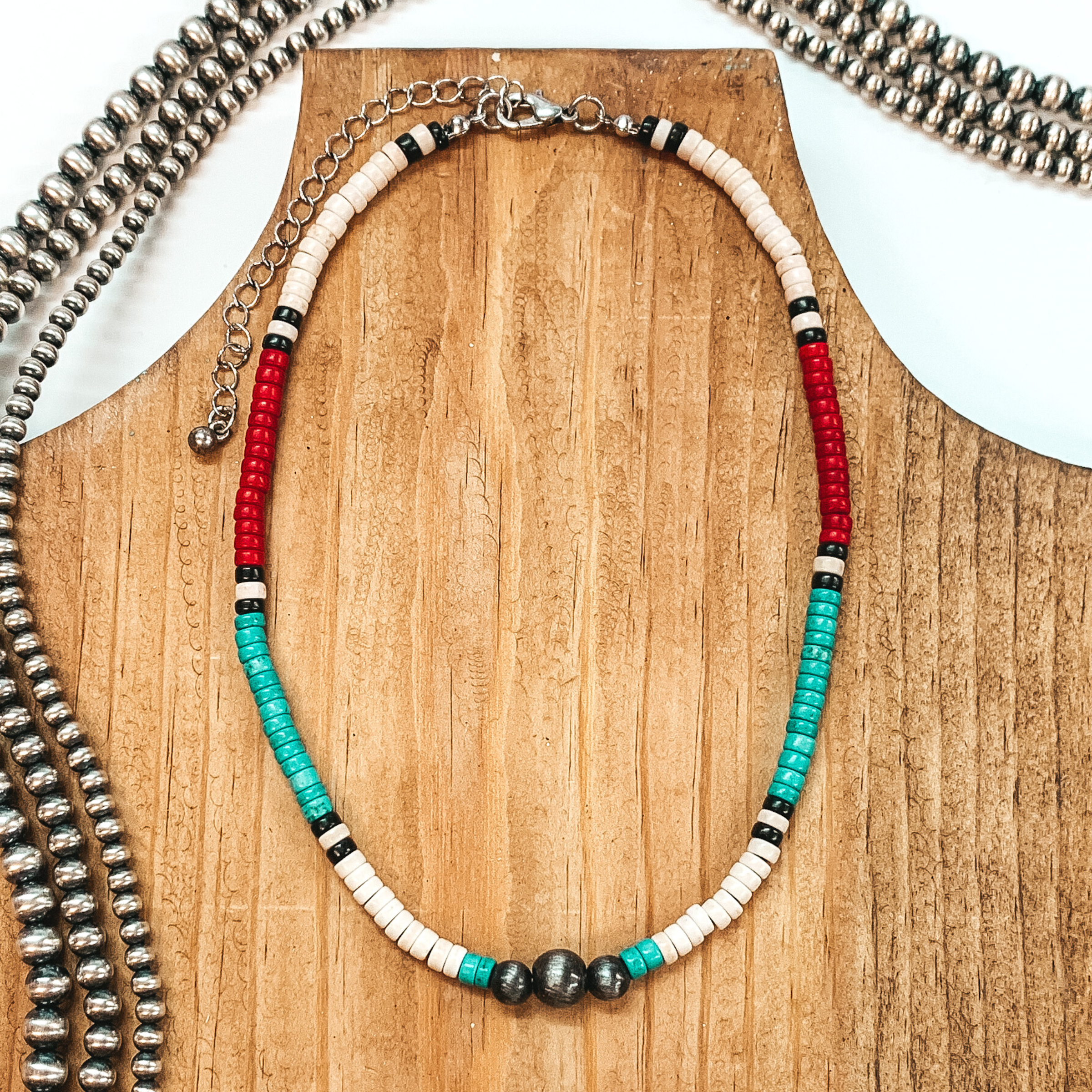 This necklace has segments of beads in the colors ivory, red, and turquoise with black beaded spacers. This necklace also includes three silver beads at the center. this necklace is pictured on a brown necklace holder on a white background with silver beads. 