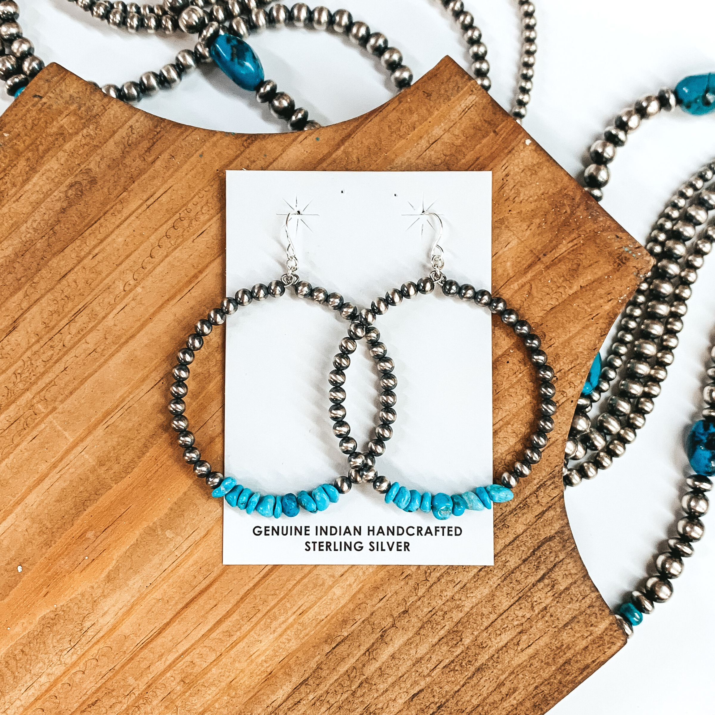 Silver fish hook earrings with a hanging silver beaded circle pendant that includes a section of turquoise stones. These earrings are pictured on a white card holder in a brown block. This is all pictured on a white background with silver beads.