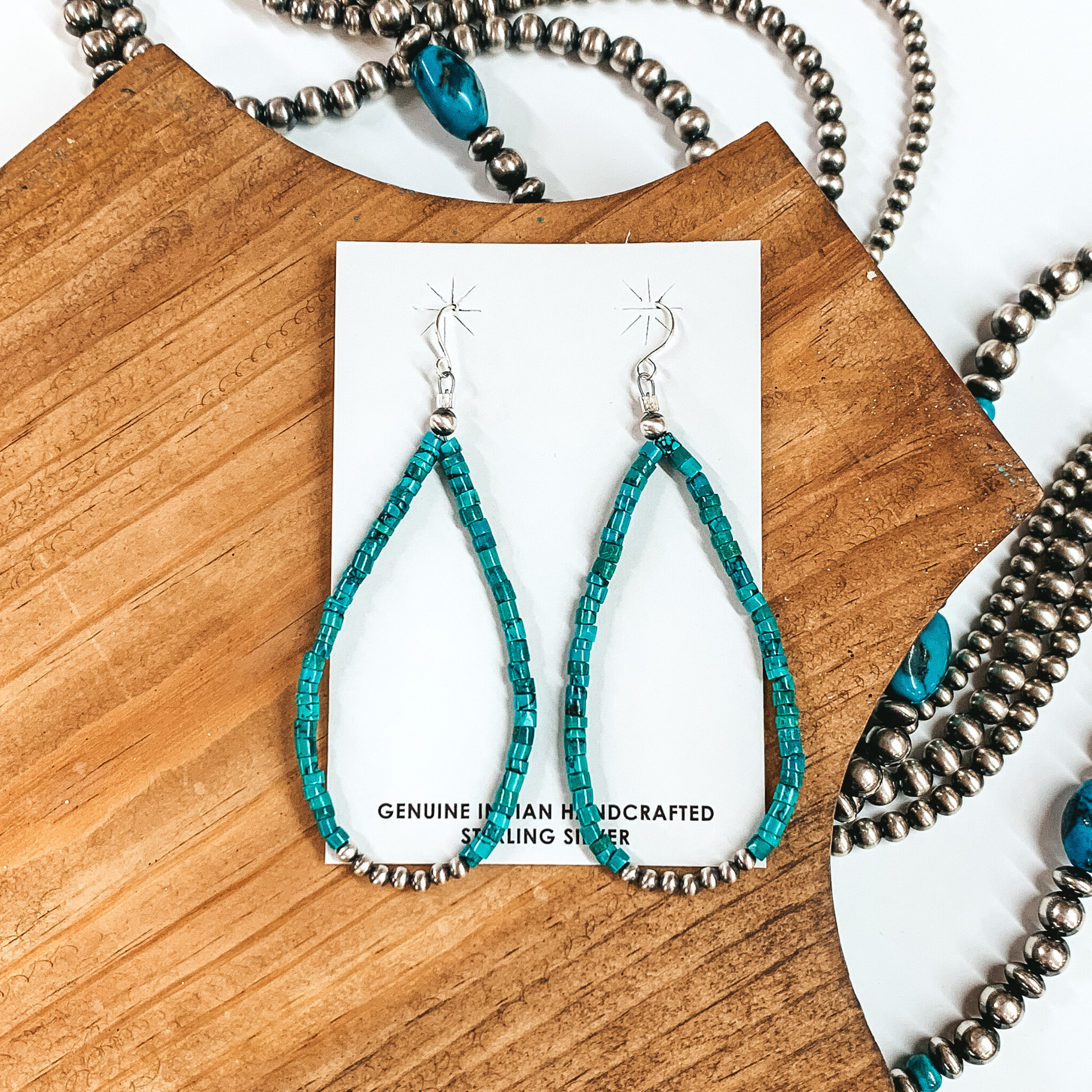 Silver fish hook earrings with a hanging teardrop pendant. This pendant is turquoise beaded with silver beads at the bottom of the teardrop. These earrings are pictured on a white card holder on top of a brown block. All of this is pictured on a white background that includes silver beaded strands. 