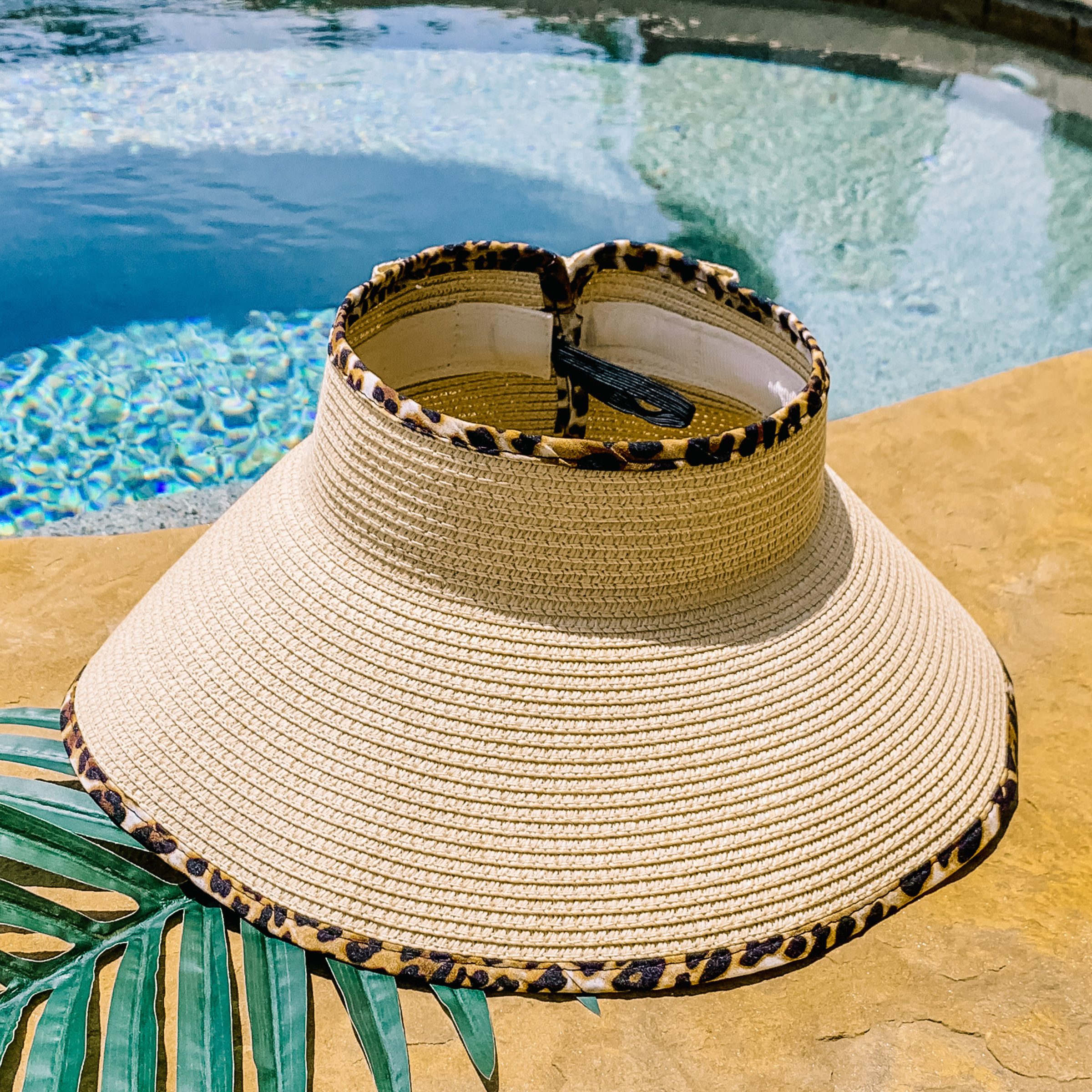 Natural, woven velcro visor with leopard print trim that is pictured with a green leaf under it. This visor is pictured on a a tan rock in front of a pool.