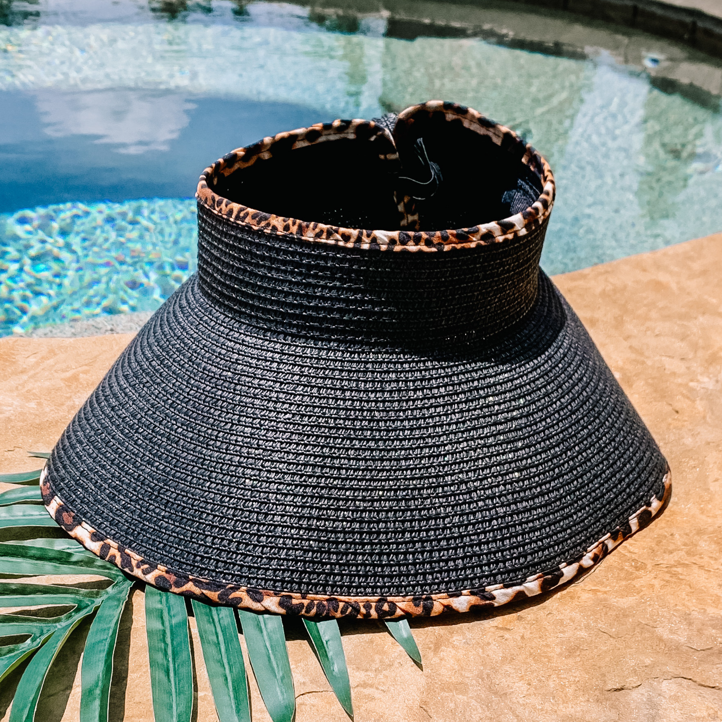 Black, woven velcro visor with leopard print trim that is pictured with a green leaf under it. This visor is pictured on a a tan rock in front of a pool.