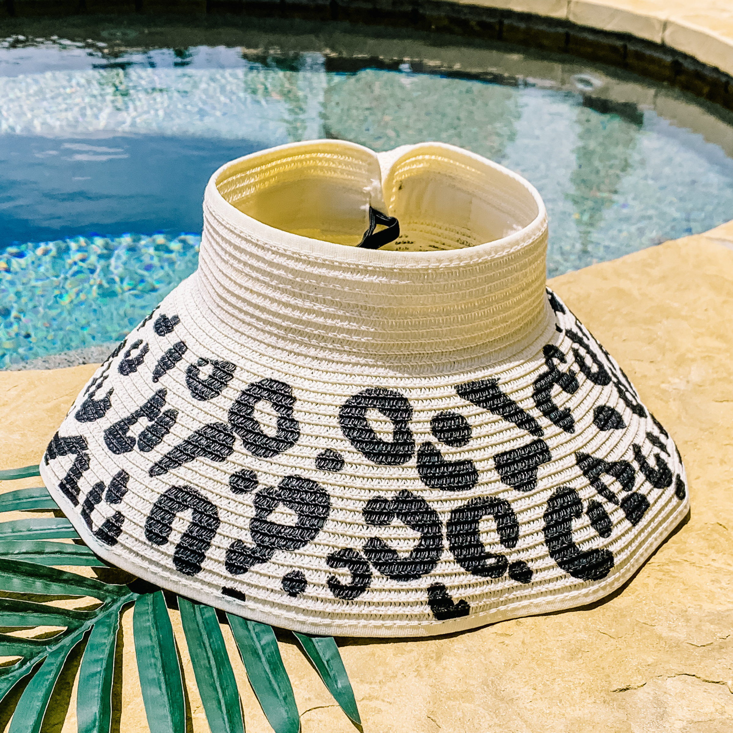 Ivory, woven velcro visor with a leopard print that is pictured with a green leaf under it. This visor is pictured on a a tan rock in front of a pool.