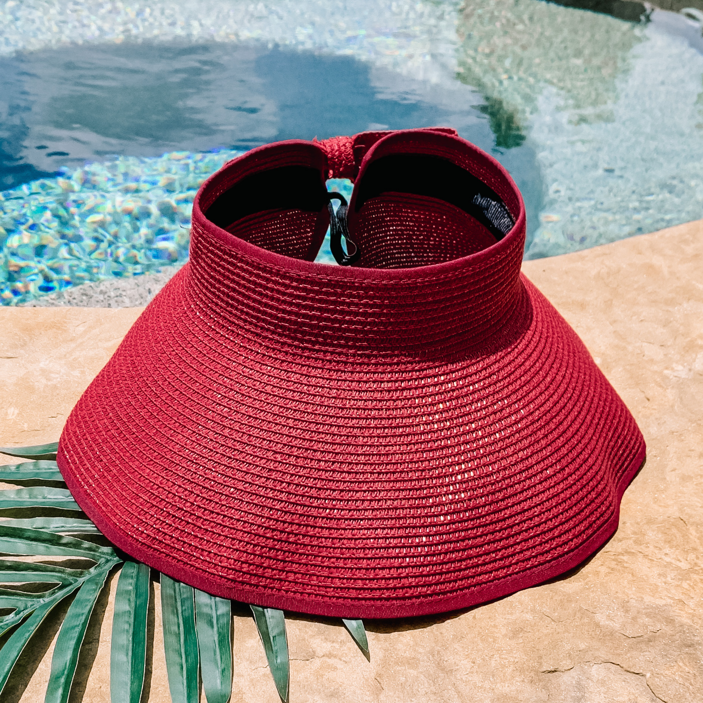 Red, woven velcro visor that is pictured with a green leaf under it. This visor is pictured on a a tan rock in front of a pool.