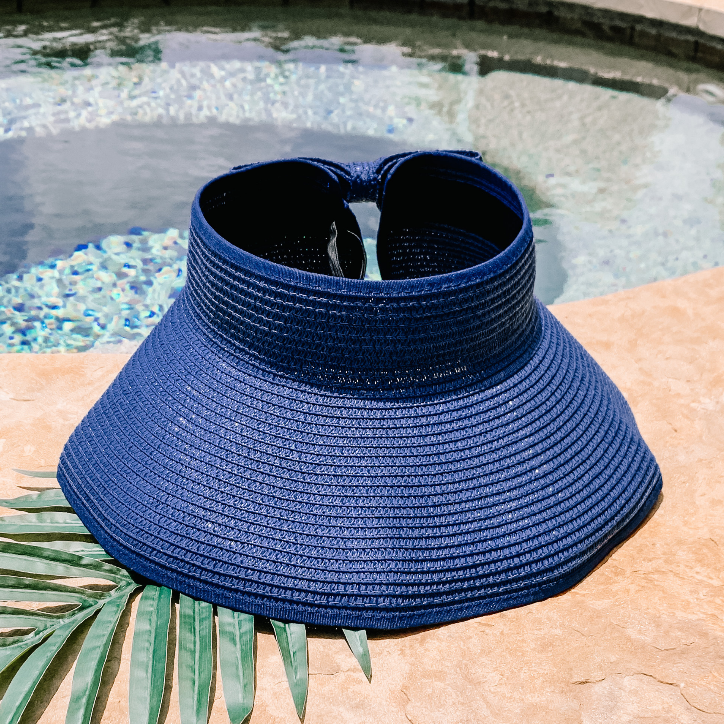 Navy blue, woven velcro visor that is pictured with a green leaf under it. This visor is pictured on a a tan rock in front of a pool.