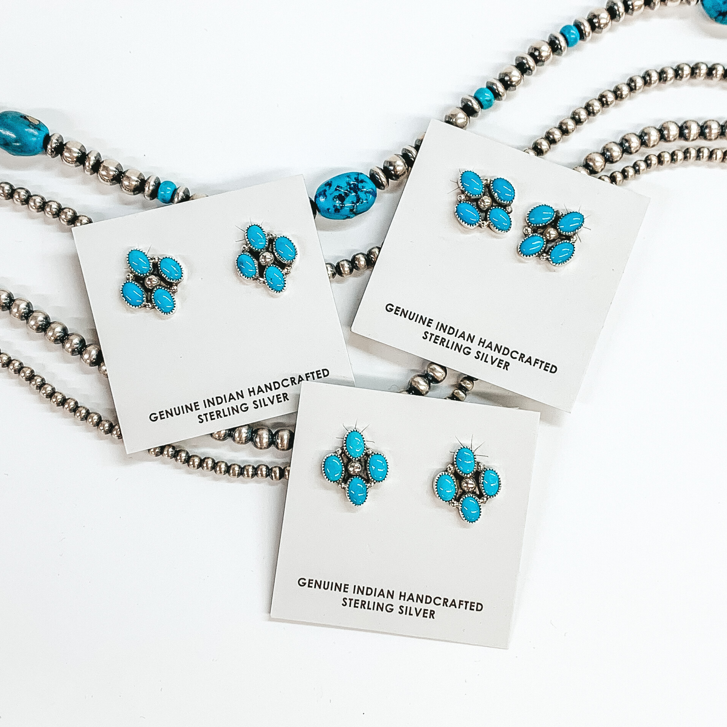 These earrings have a silver base and center silver bead with four oval turquoise stones. These earrings are pictured on a white cardstock pictured on a white background with silver beads under them. 