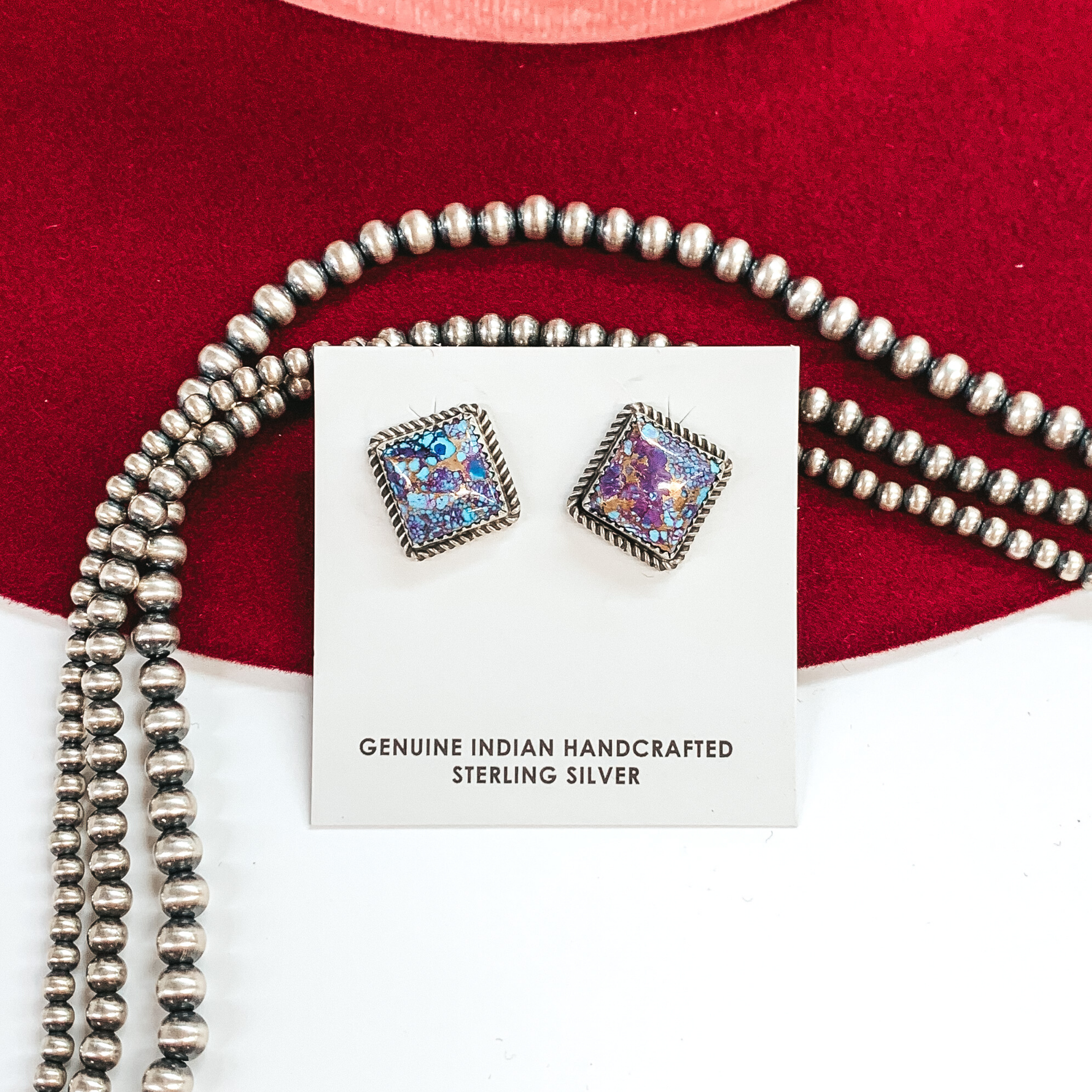 Silver square stud earrings with purple mojave  and turquoise stone inlay. these earrings are pictured on a white cardstock on a white and red background with silver beads behind them. 