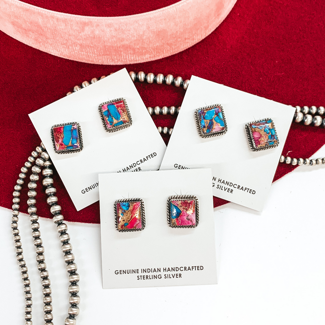 Silver square stud earrings with purple dahlia remix stone inlay. These earrings are pictured on white cardstock and on a white and red background with silver beads under.