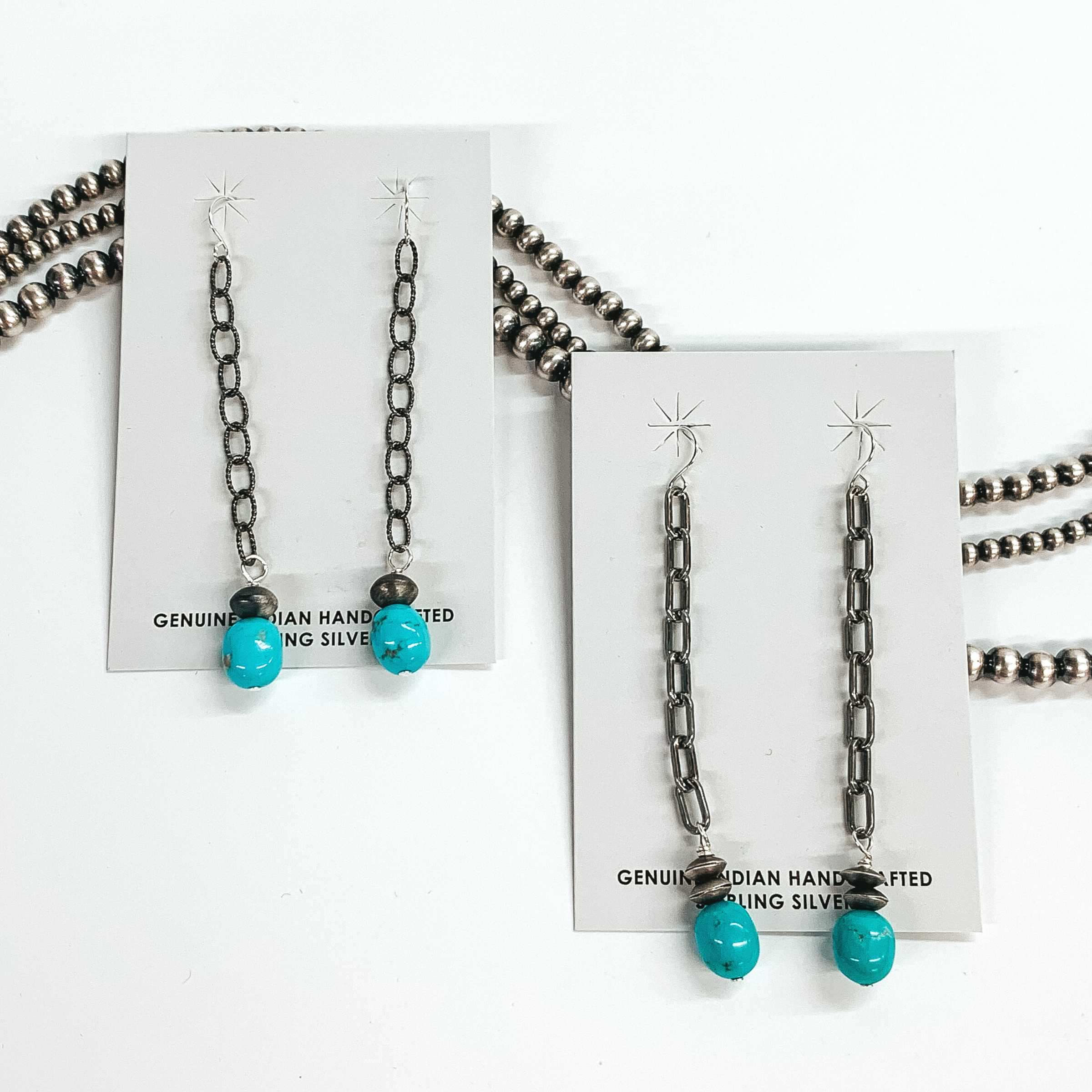 Silver fish hook earrings with a silver chain drop that has a Navajo pearl charm and turquoise stone at the bottom of the chain. These earrings are pictured on white cardstock on a white background with silver beads behind them. 