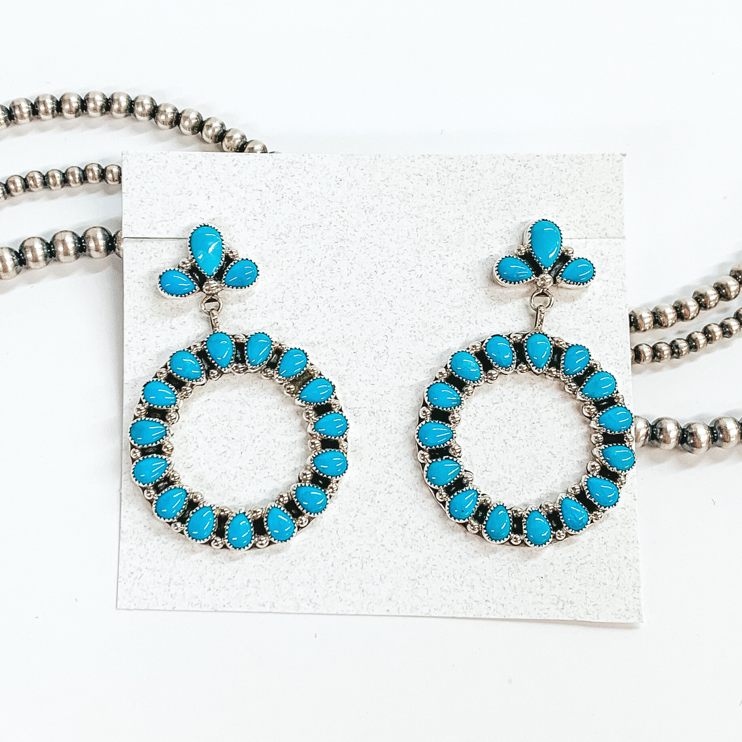 Silver circle drop earrings. The studs have three turquoise stones while the circle has a turquoise stone inlay. These earrings are pictured on a white cardstock on a white background with silver beads behind them. 