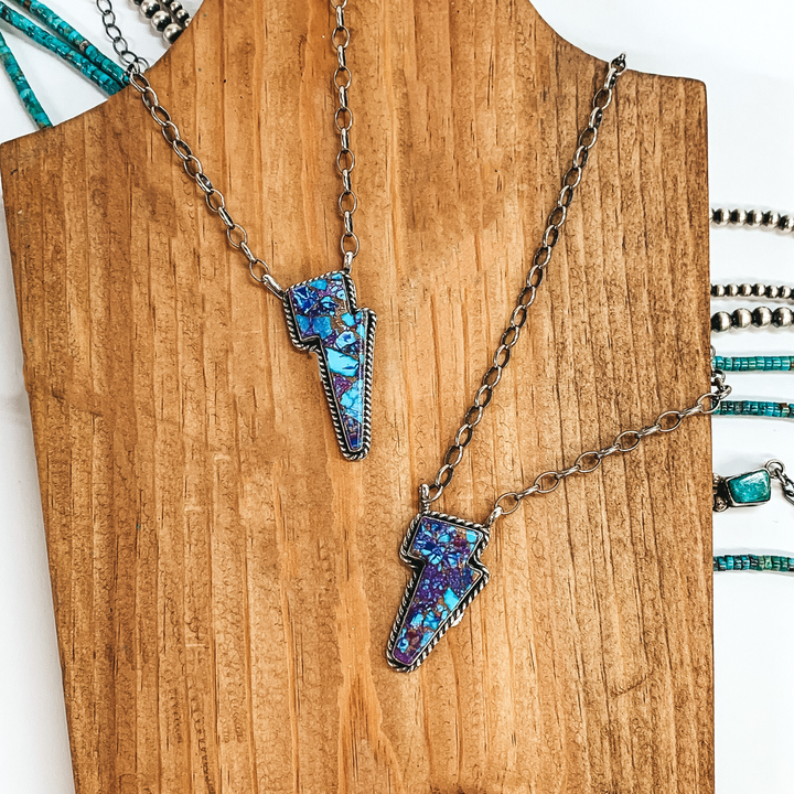 Two silver chained necklaces that have lightning bolt pendants that have purple mojave stones. These necklaces are on a pictured on a brown block on a white background. 