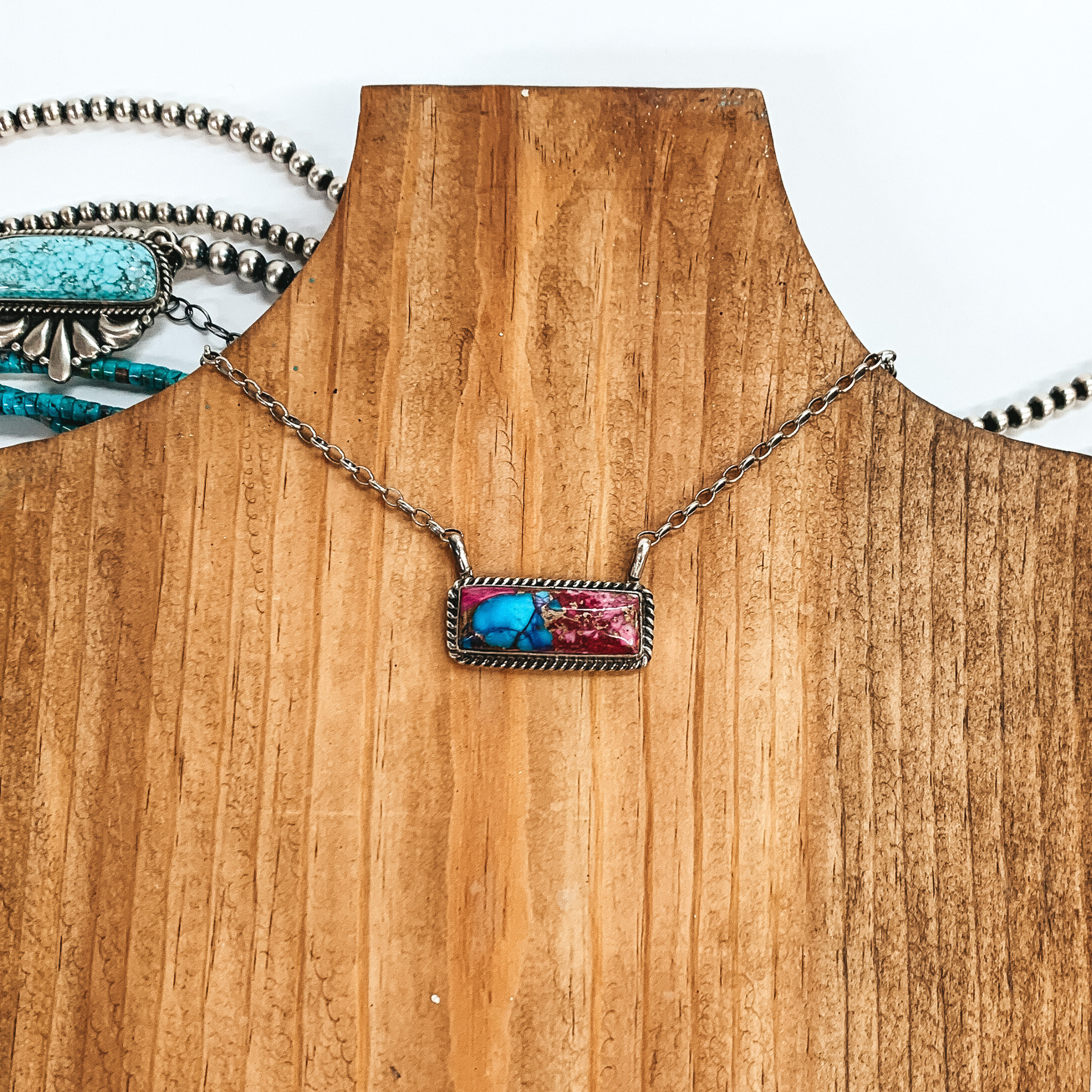 This necklace is a silver chain necklace with a rectangle dhalia remix stone pendant with silver rope detailing. This necklace is pictured on a brown necklace holder on a white background. 