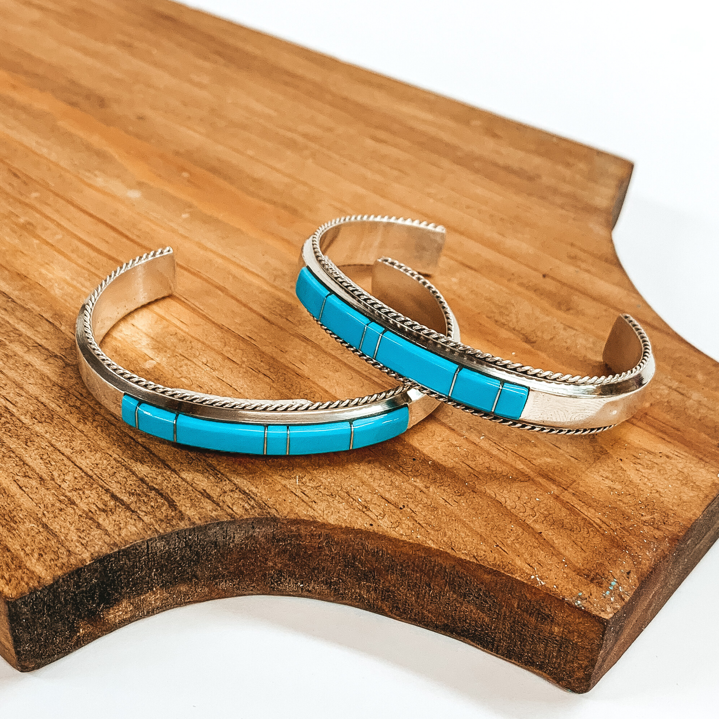 Two silver cuff bracelet with rope detailing outline. These bracelets include turquoise stones divided by silver spacers. These bracelets are pictured on top of a brown block on a white background. 