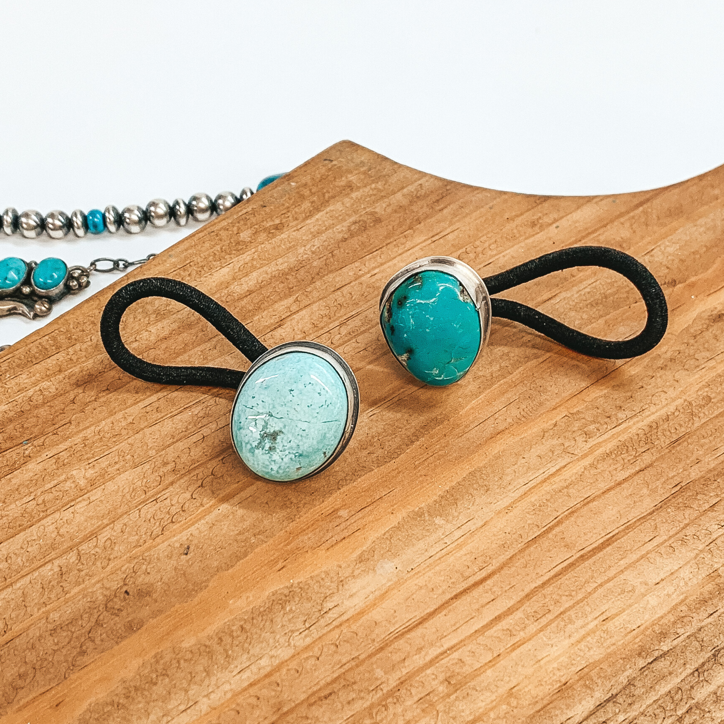 Asymmetrical turquoise stones set in silver with a black hair tie connected to the back. These hair ties are pictured on a brown block on a white background.  