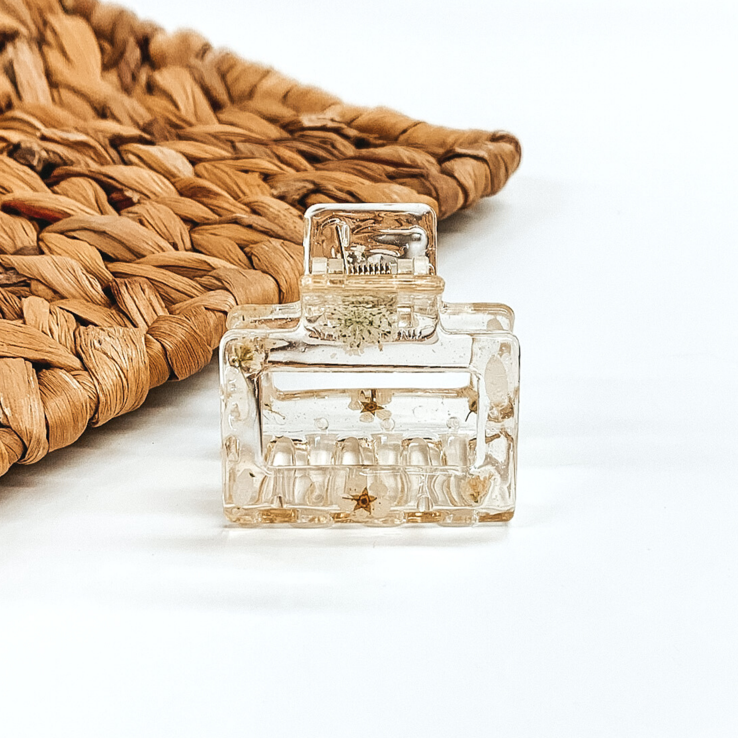 Clear square clip, that has ivory pressed flowers inside the clip. This clip is pictured on a white background with basket weave behind it. 
