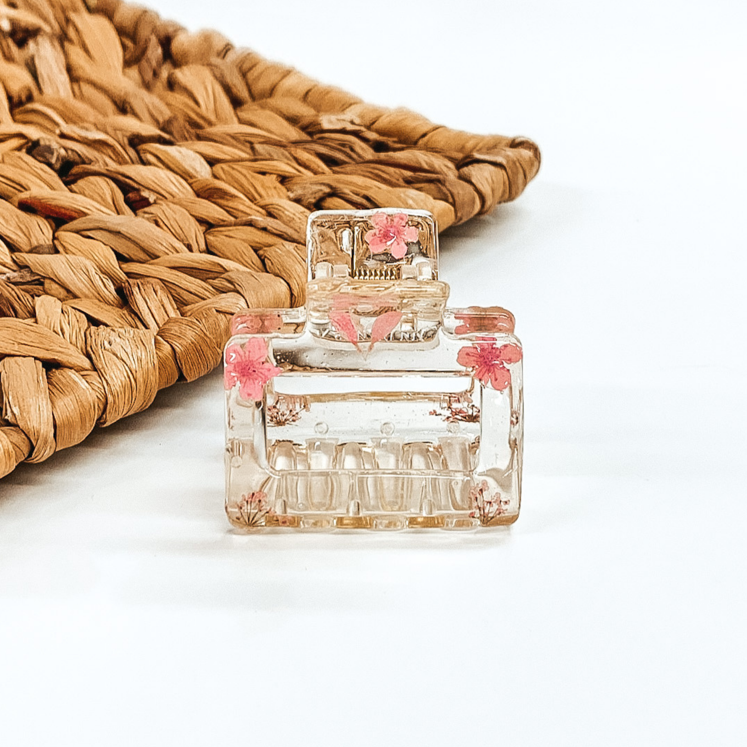 Clear square clip, that has light pink pressed flowers inside the clip. This clip is pictured on a white background with basket weave behind it. 