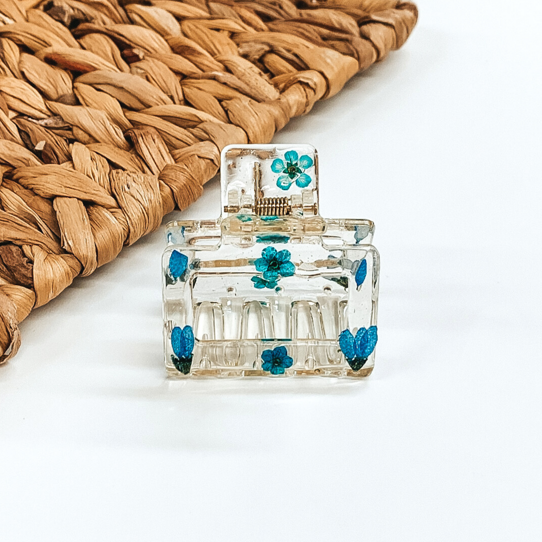 Clear square clip, that has blue pressed flowers inside the clip. This clip is pictured on a white background with basket weave behind it. 