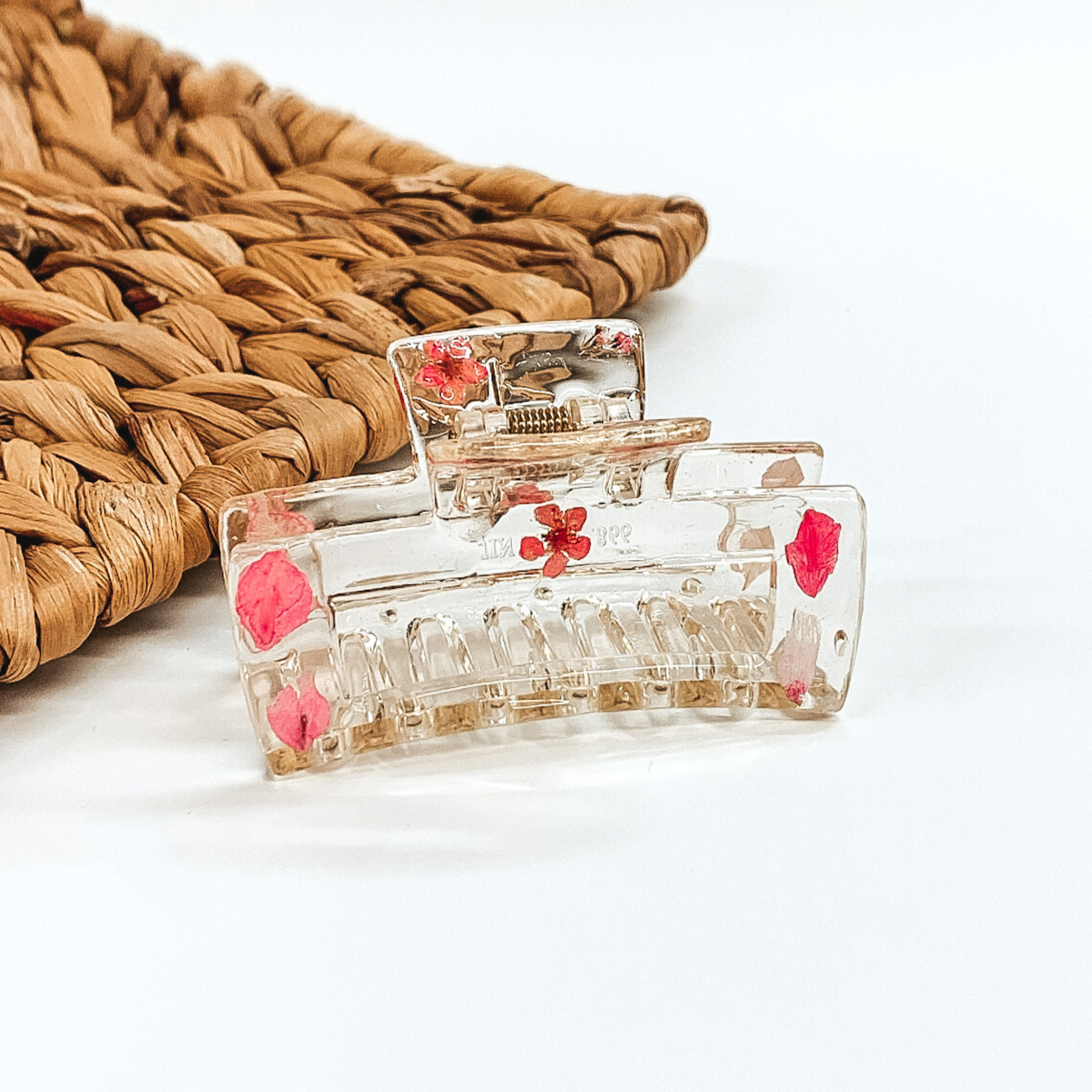 Clear rectangle clip, that has pink pressed flowers inside the clip. This clip is pictured on a white background with basket weave behind it. 