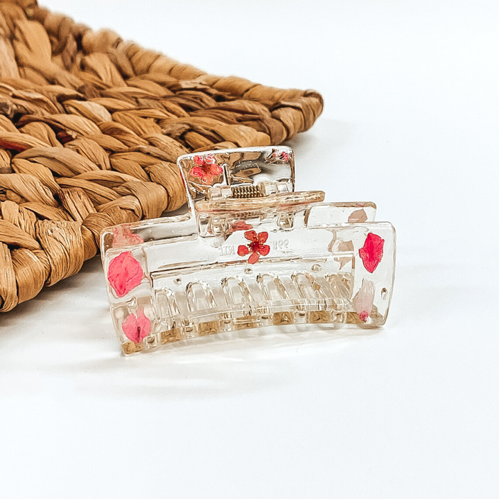Clear rectangle clip, that has pink pressed flowers inside the clip. This clip is pictured on a white background with basket weave behind it. 