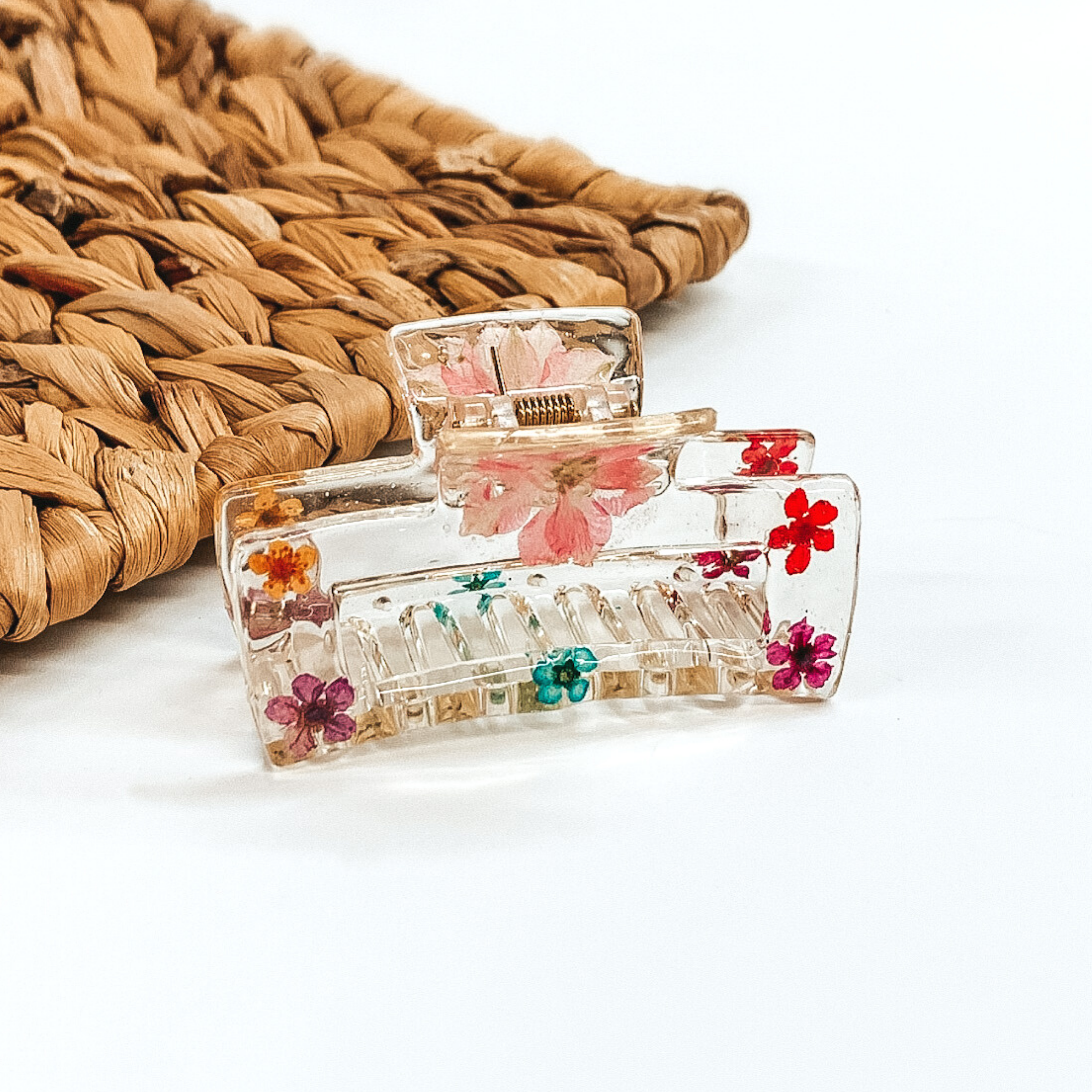 Clear rectangle clip, that has multicolored pressed flowers inside the clip. This clip is pictured on a white background with basket weave behind it.