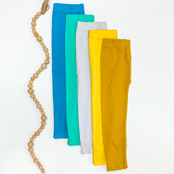 Leggings are pictured folded in half laying on top of each other on a white background. The leggings pictured ar ein colors turquoise, mint, lavender, bright yellow, and mustard with tan beads to the left of the stack.