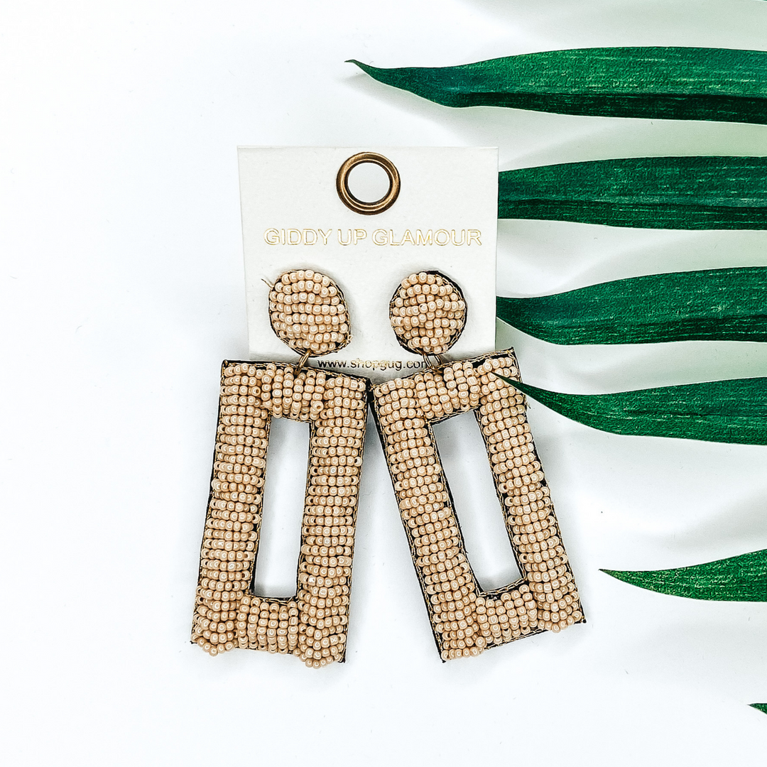 Ivory beaded circle post back earrings with a hanging beaded open rectangle pendant. This pair of earrings is pictured on a white background with green leaves behind them.