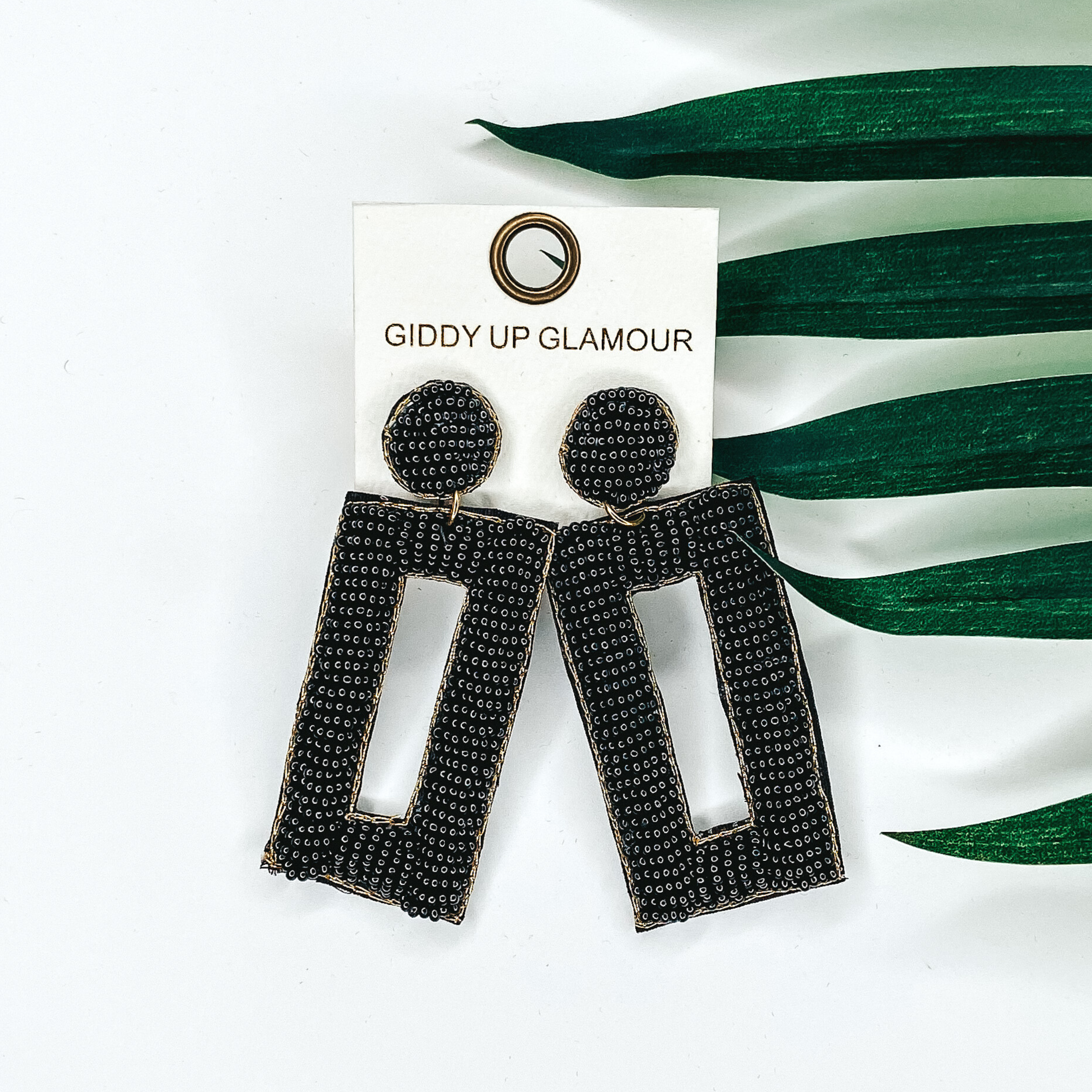 Black beaded circle post back earrings with a hanging beaded open rectangle pendant. This pair of earrings is pictured on a white background with green leaves behind them. 