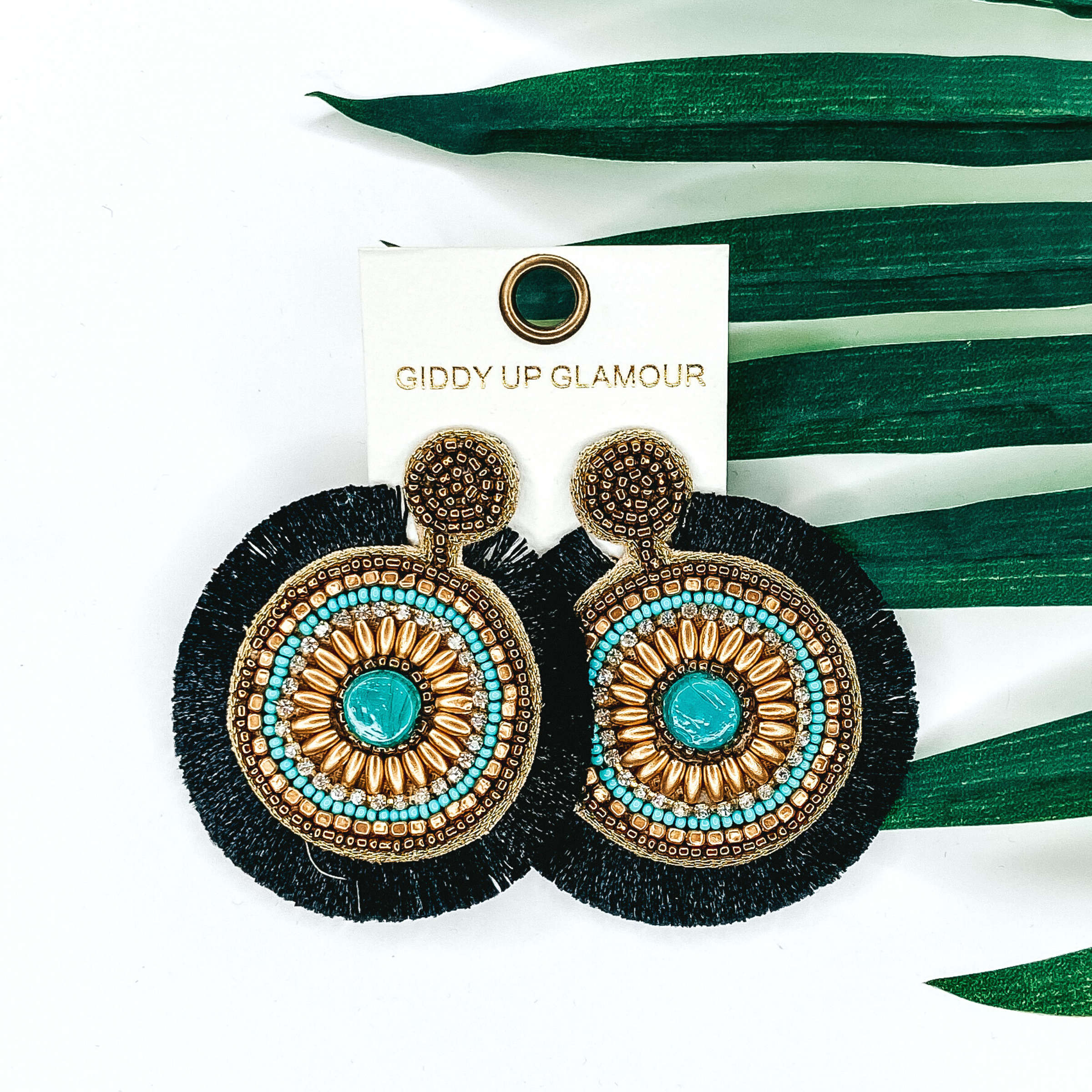 Circle beaded earrings with a black fringe out line. These earrings include gold and turquoise beads. These earrings are pictured on a white background with green leaves behind them. 