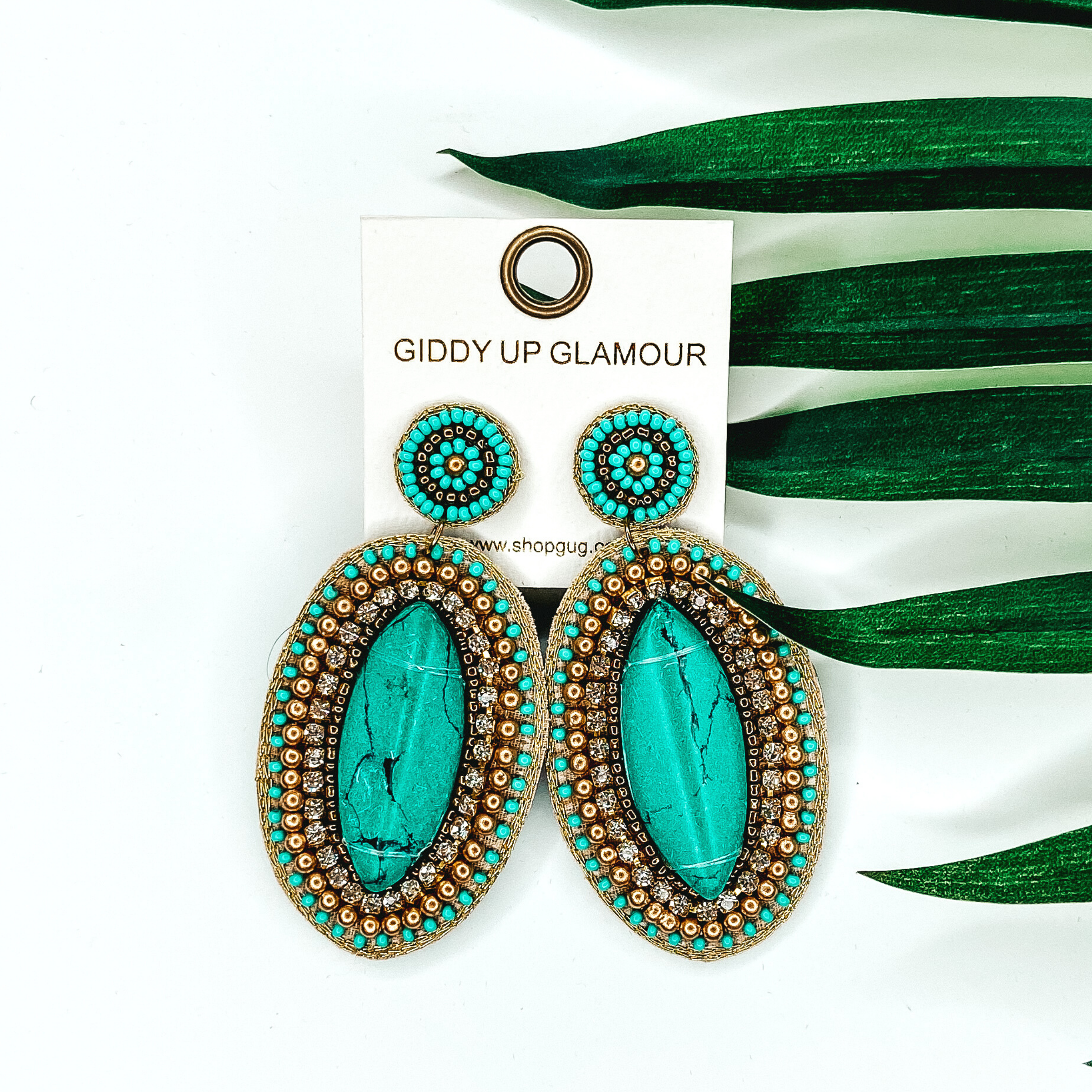Beaded oval earrings in gold and turquoise. These earrings include a big center turquoise stone. These earrings are pictured on a white background with green leaves behind them.