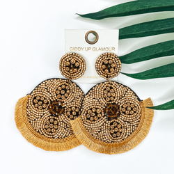 Gold beaded circle post back earrings. Hanging from the earrings are black beaded circle pendant with black fringe. These earrings are pictured on a white background with green leaves behind them.