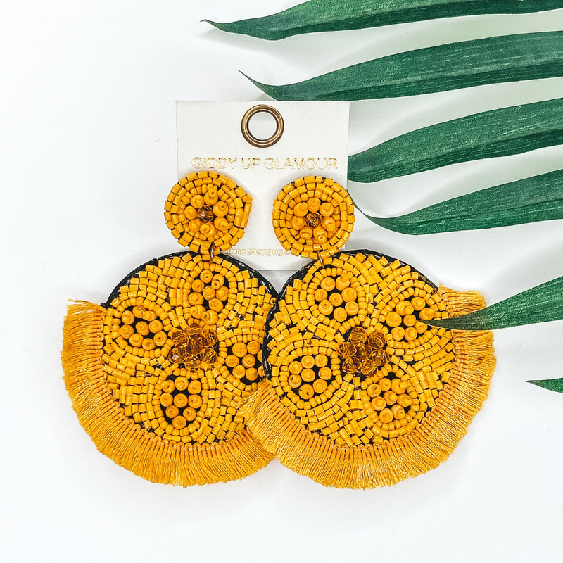 Yellow beaded circle post back earrings. Hanging from the earrings are black beaded circle pendant with black fringe. These earrings are pictured on a white background with green leaves behind them.