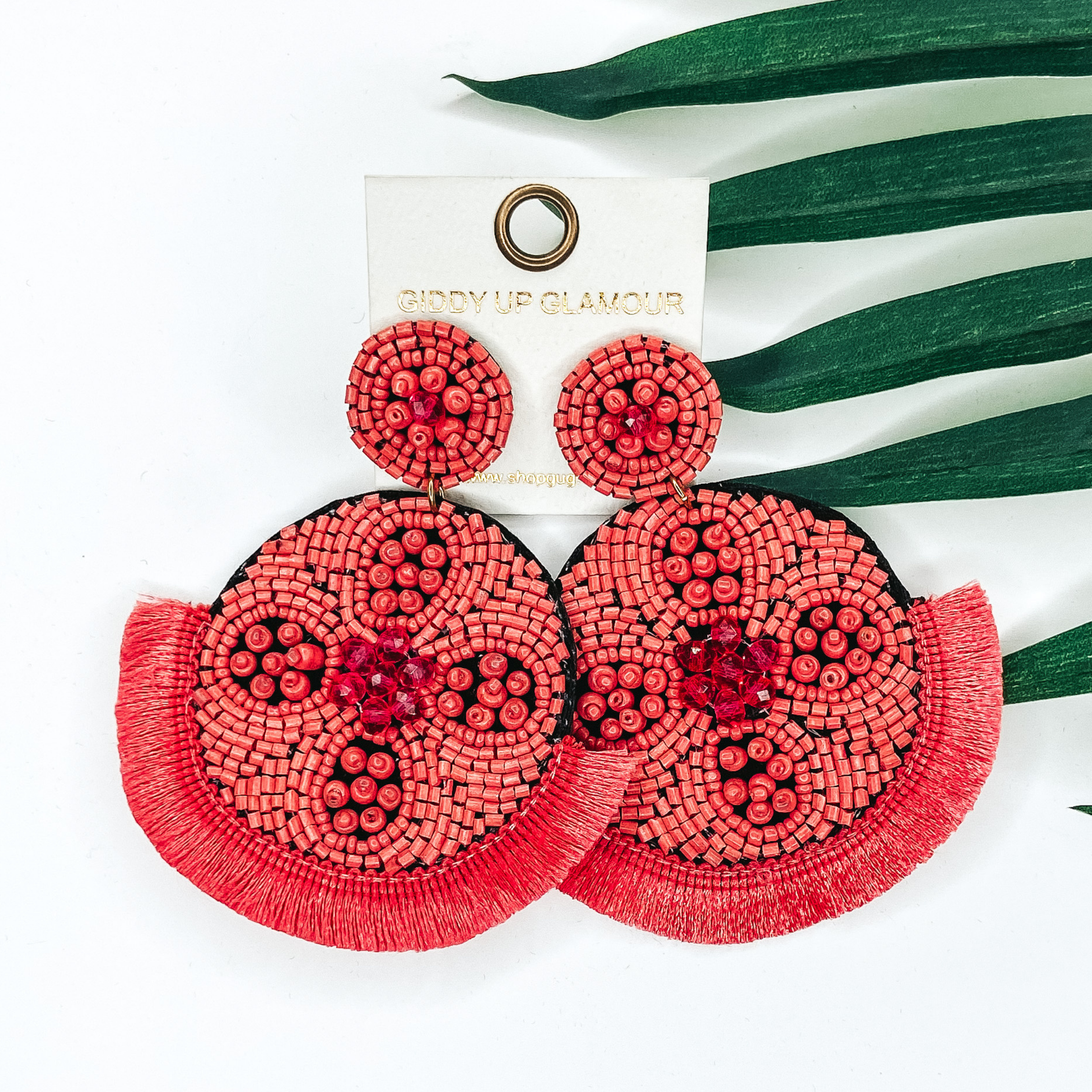 Coral beaded circle post back earrings. Hanging from the earrings are black beaded circle pendant with black fringe. These earrings are pictured on a white background with green leaves behind them.