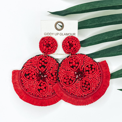 Red beaded circle post back earrings. Hanging from the earrings are black beaded circle pendant with black fringe. These earrings are pictured on a white background with green leaves behind them.