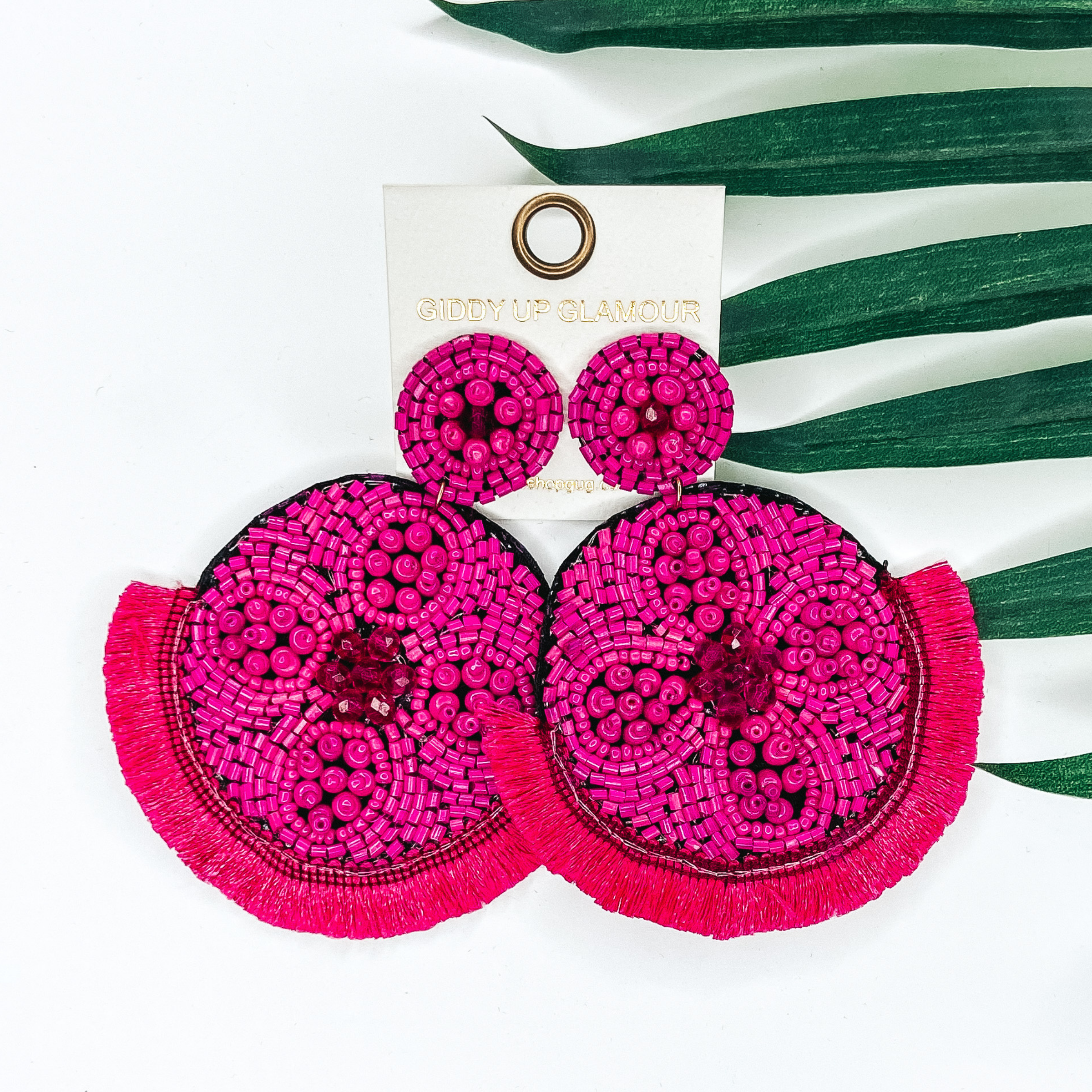 Fuchsia beaded circle post back earrings. Hanging from the earrings are black beaded circle pendant with black fringe. These earrings are pictured on a white background with green leaves behind them.