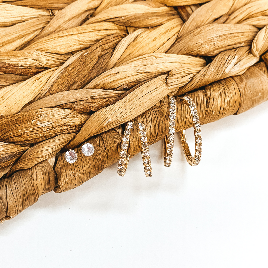 Clear crystal studs with two pairs of gold hoops with clear crystals that are different sizes. these earrings are pictured on a white background up against a tan basket weave. 