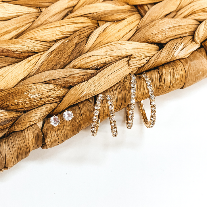 Clear crystal studs with two pairs of gold hoops with clear crystals that are different sizes. these earrings are pictured on a white background up against a tan basket weave. 
