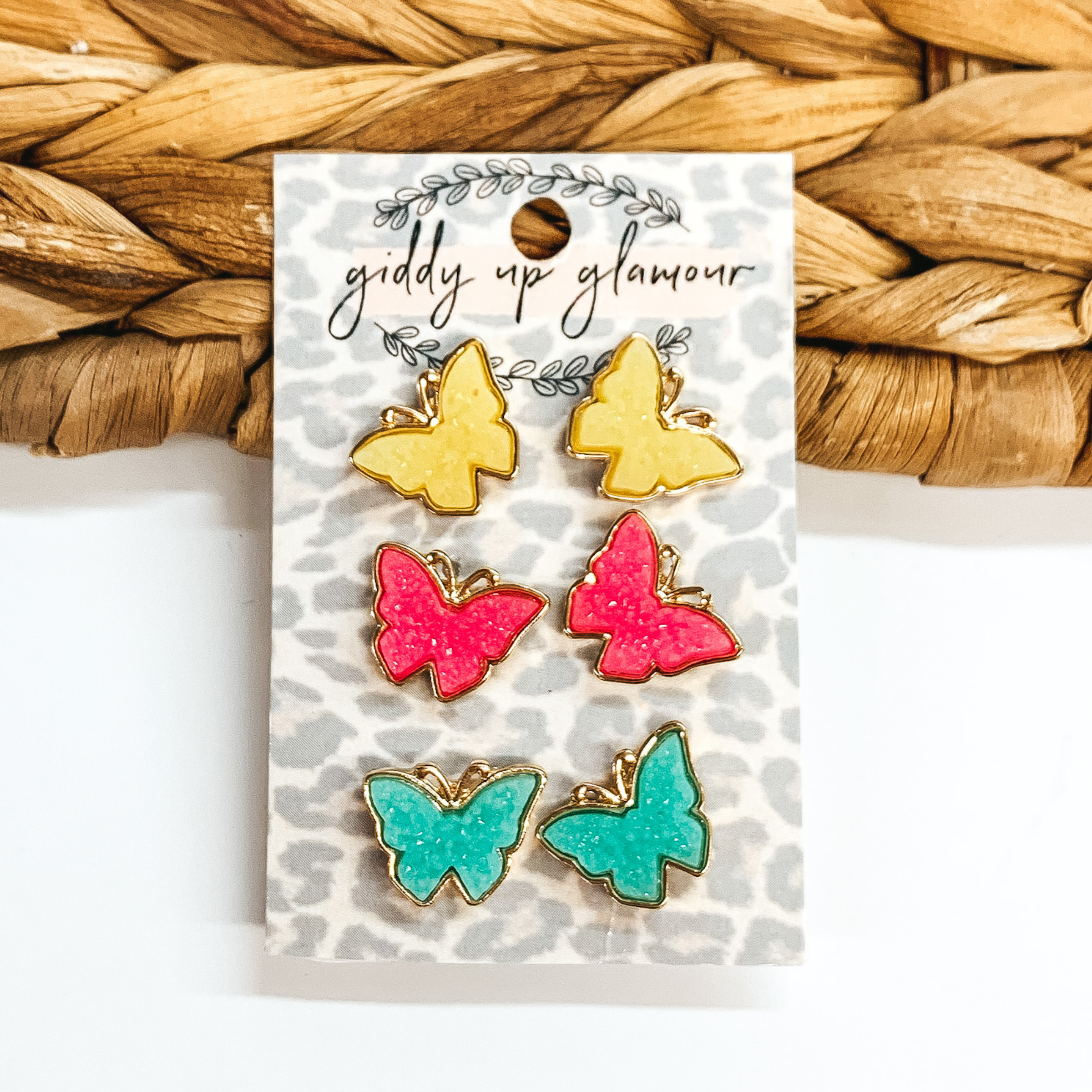 Set of three butterfly earrings with druzy stones in colors, neon yellow, hot pink, and teal. These earrings are pictured on a leopard card holder on a white and basket weave background. 