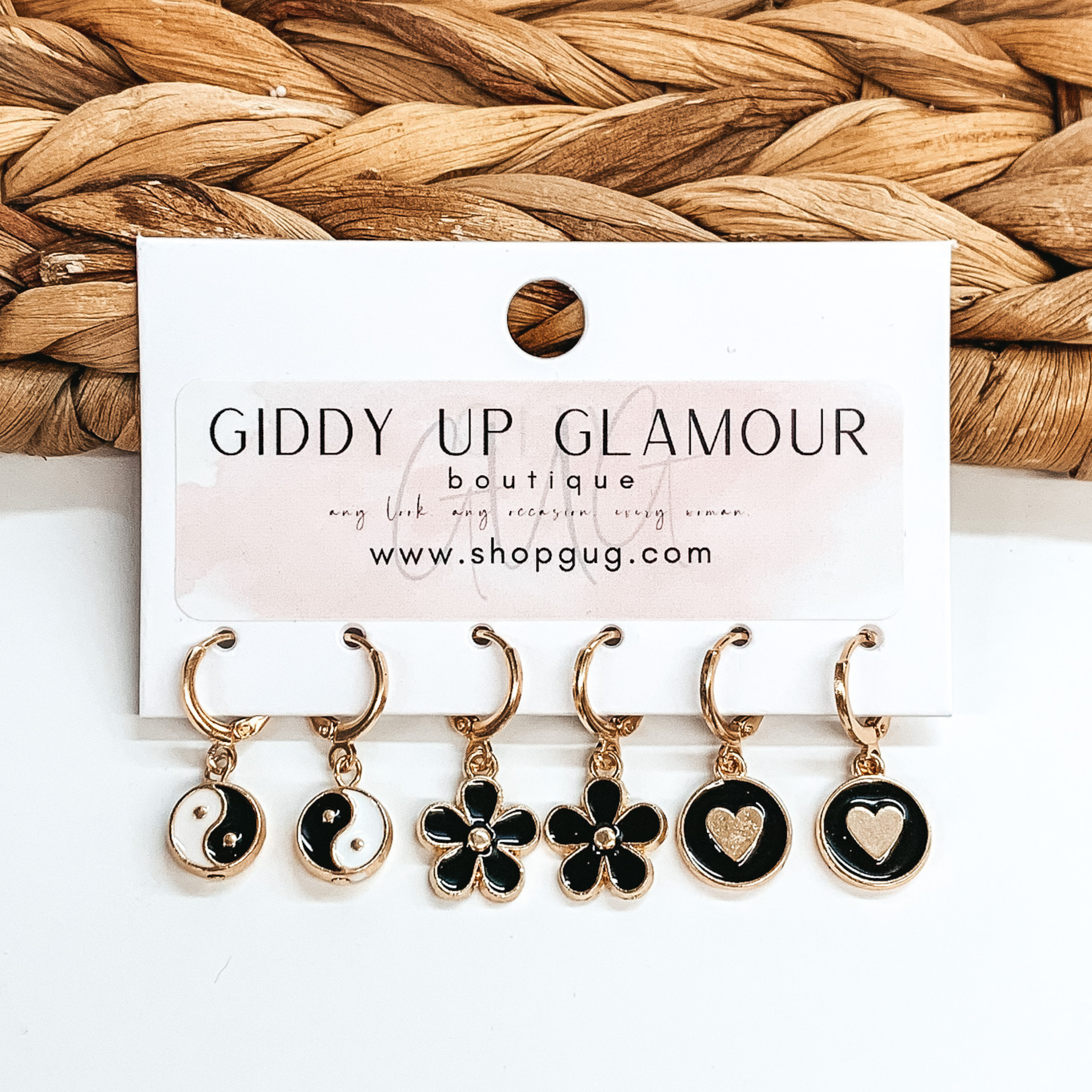 Set of three gold hoop earrings each with a different pendant. The pendants include black and white ying and yang circle pendants, black flower pendants, and black circle pendants with gold hearts in the center. These earrings are pictured on a white card holder on a white background with basket weave in the background. 