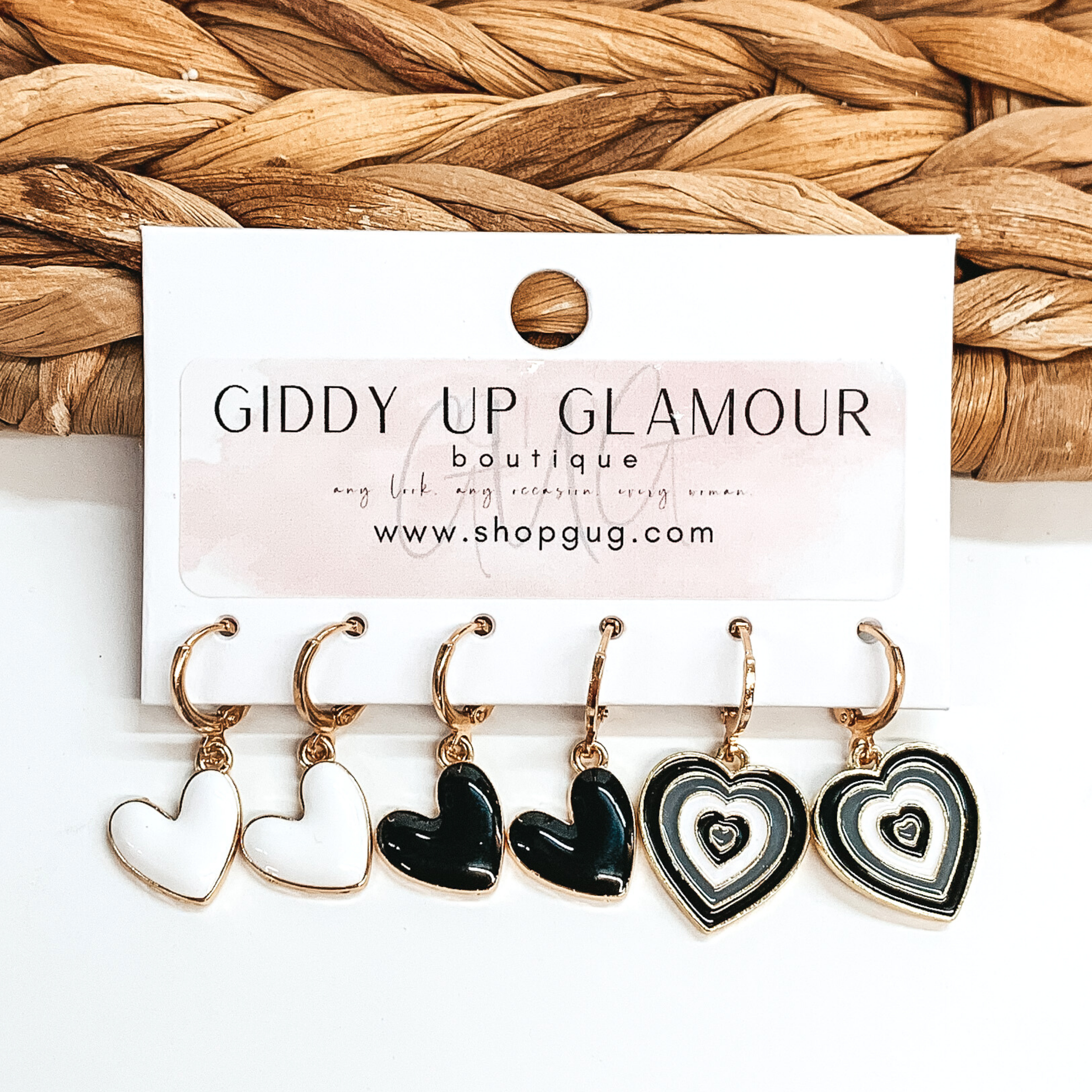 Set of three gold hoop earrings that have heart pendants hanging. the pendants include white hearts, black hearts, and heart pendants that have layers in black and white. These earrings are pictured on a white card holder on a white background with basket weave in the background.