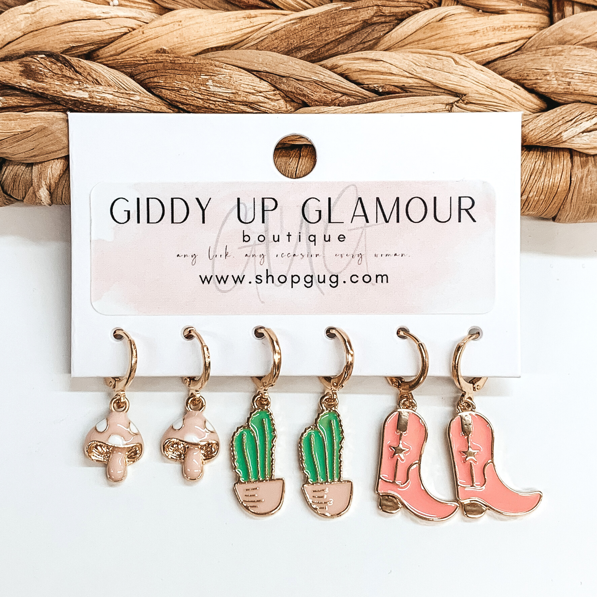 Set of three gold hoop earrings that have western pendants hanging. The pendants include light pink mushrooms, green cacti, and pink boots. These earrings are pictured on a white card holder on a white background with basket weave in the background.
