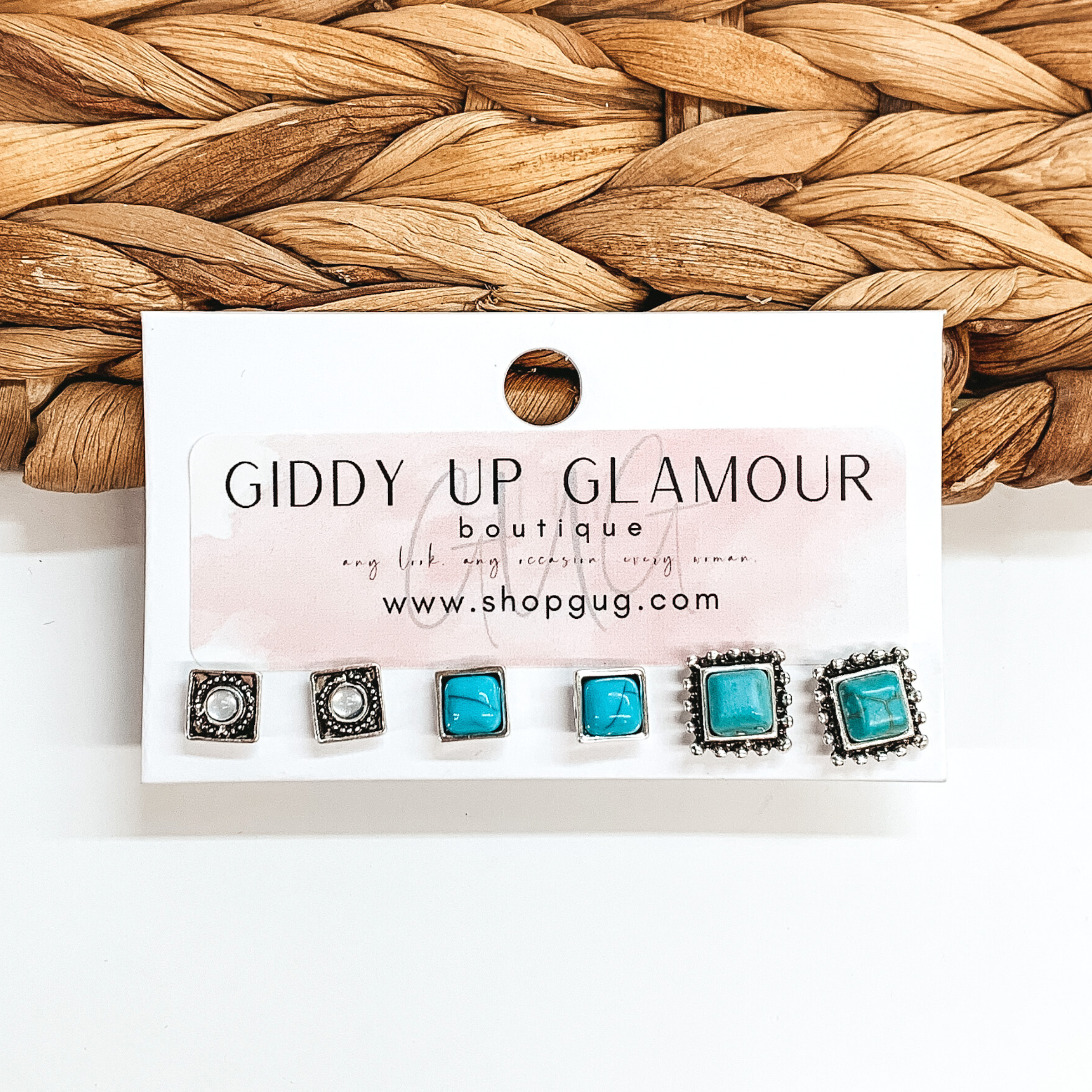 Set of three silver square stud earrings. Two pairs of studs include turquoise colored stones and the last set includes clear stones. These earrings are pictured on a white card holder on a white background with basket weave in the background.