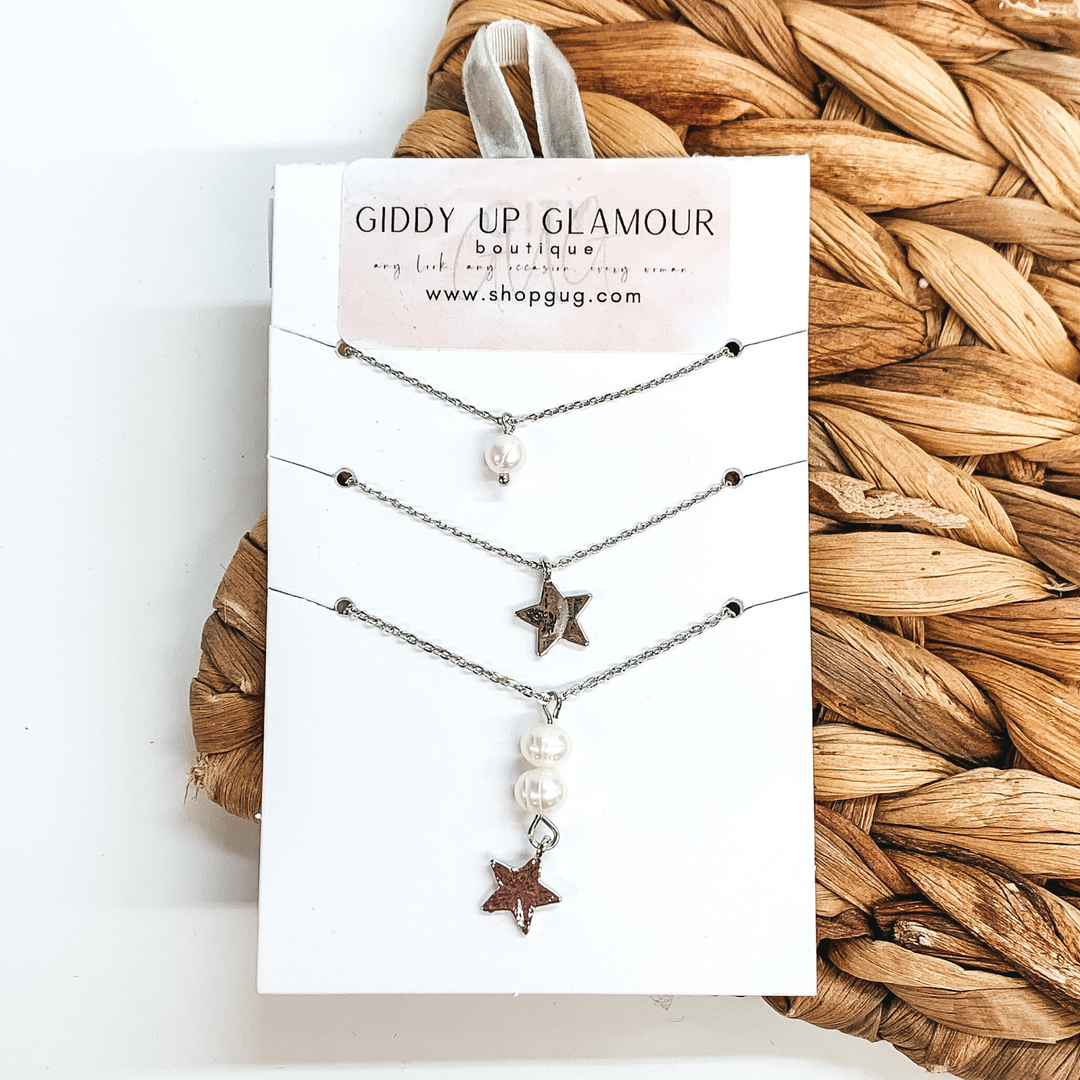 On a white card holder there is three silver chain necklaces. The first necklace has a single white pearl charm, the second necklace has a single, silver star charm, and the third necklace has two pearl charms with a hanging silver star. This is all pictured on a white and backet weave background.