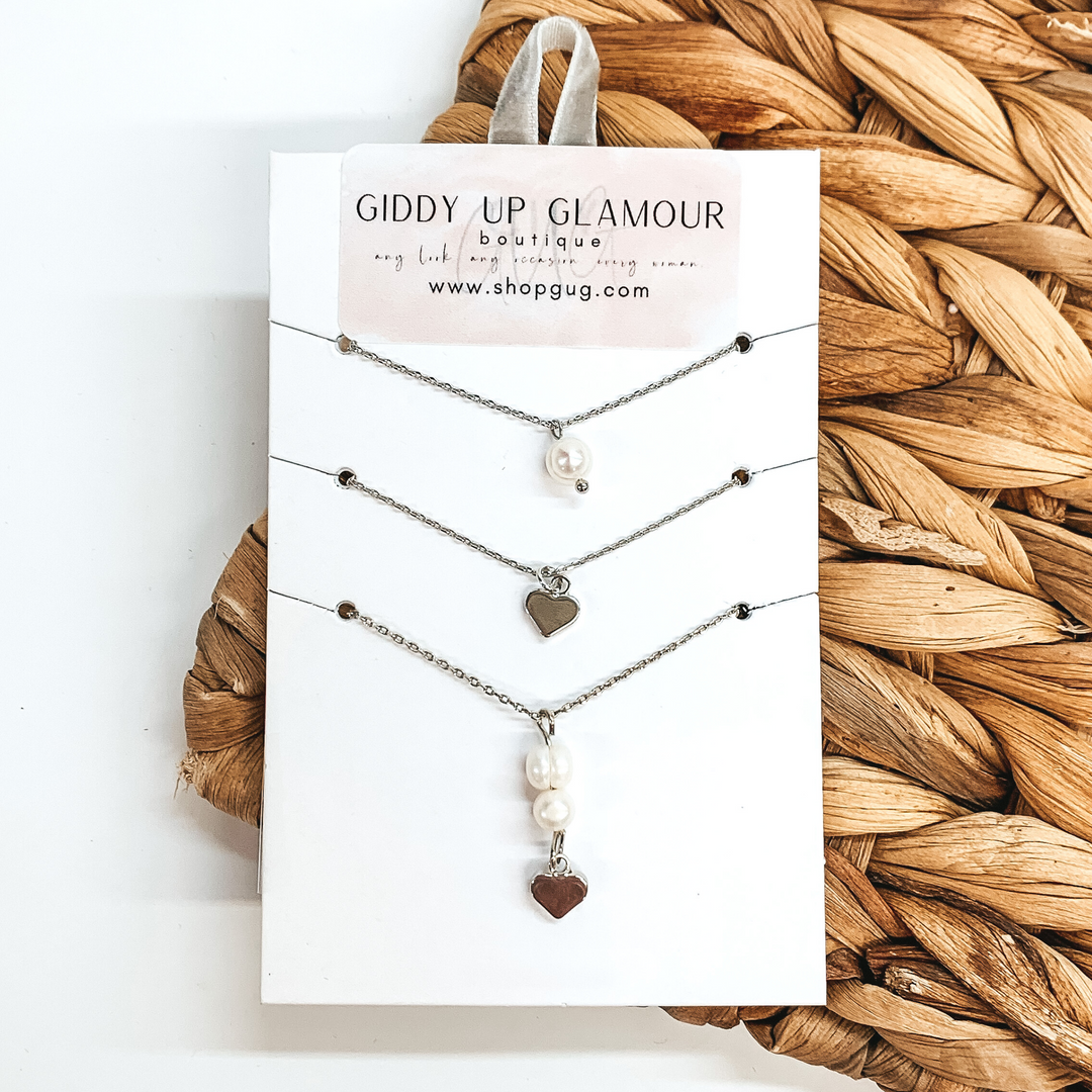 On a white card holder there is silver chain necklaces. The first necklace has a single white pearl charm, the second necklace has a single, silver heart charm, and the third necklace has two pearl charms with a hanging heart cross. This is all pictured on a white and backet weave background.