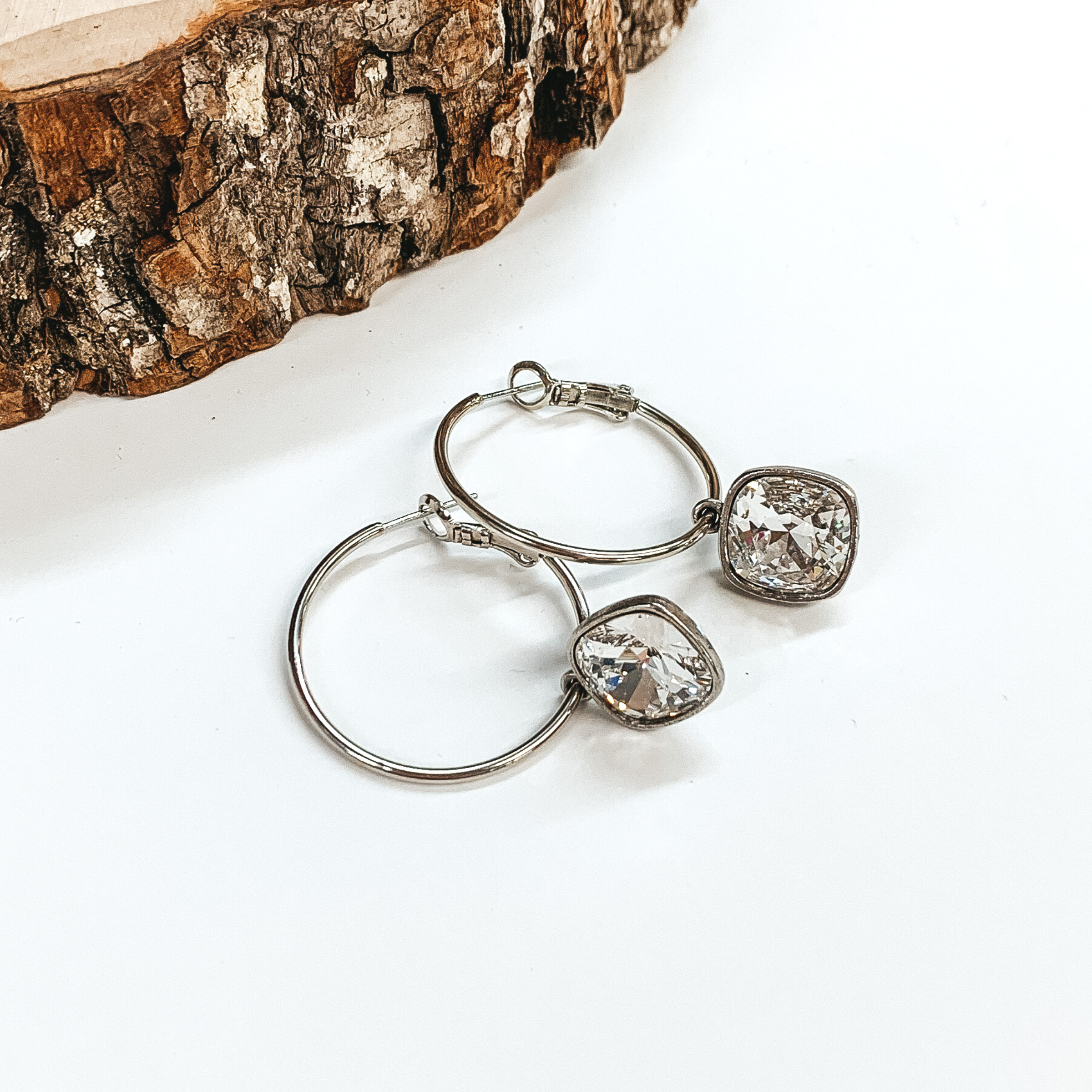 Silver hoop earrings with a clear cushion cut crystal. These earrings are pictured on a white background with a piece of wood on the top left corner. 
