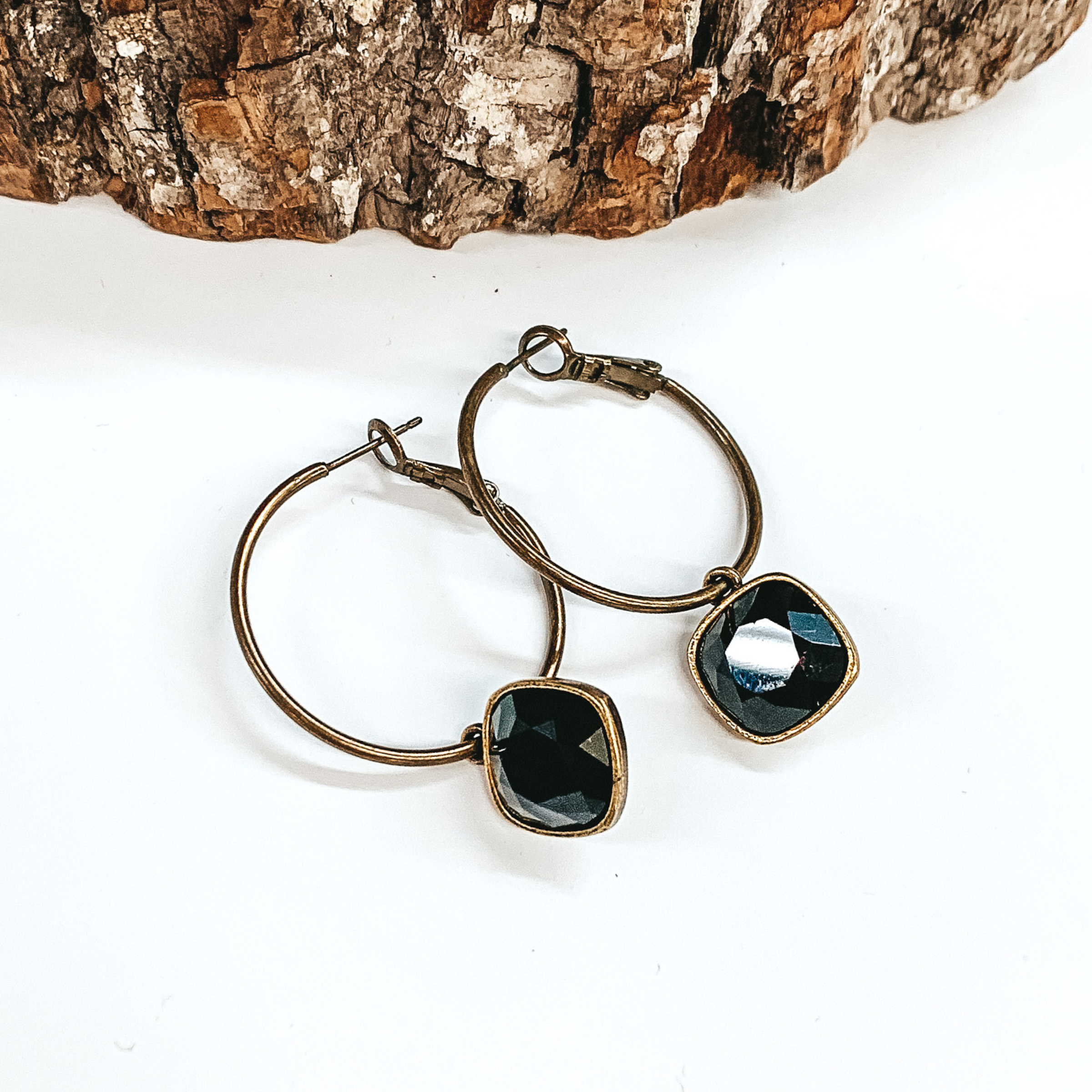 Two bronze hoop earrings with a square black crystal. These earrings are pictured on a white background with a piece of wood at the top.