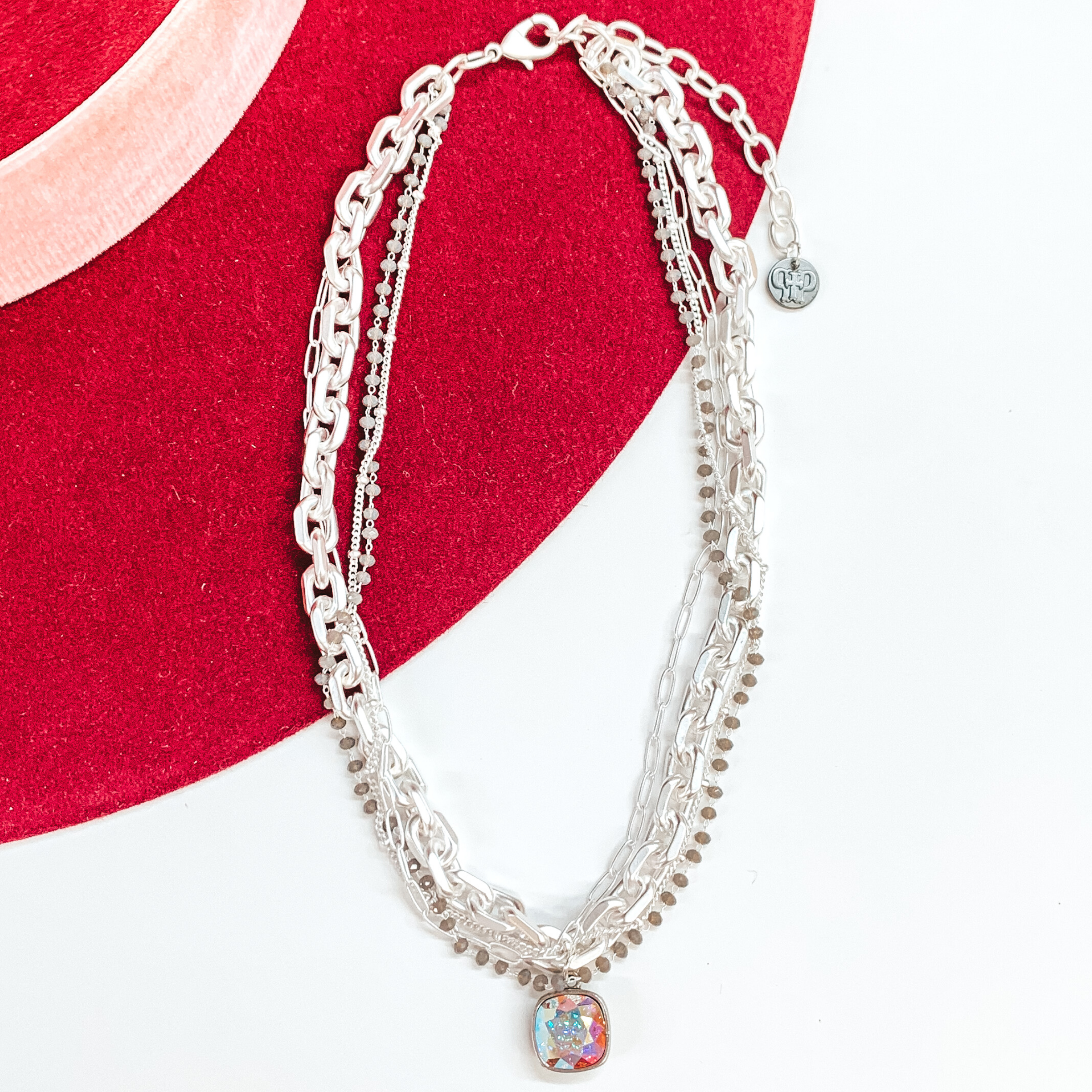 Silver, adjustable necklace that includes a thick chain strand, crystal beaded strand, a thin paperlcip chain strand, and a small chain strand. There is also an AB cushion cut crystal drop. This necklace is pictured on a white and red background.