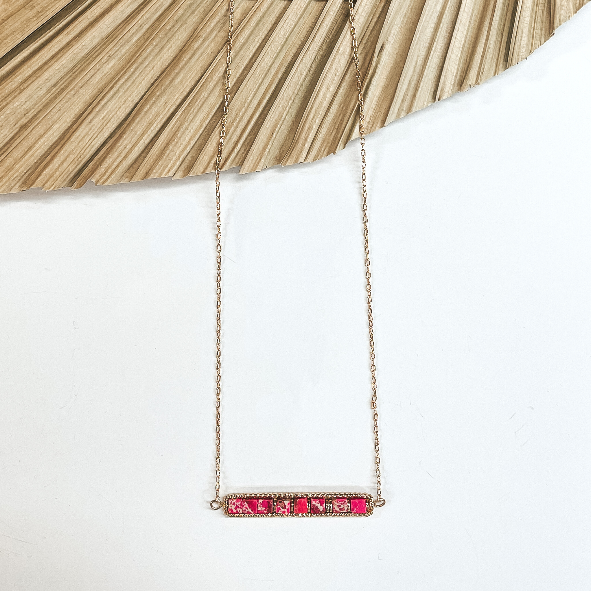 This is a gold necklace with a horizontal bar  pendant with semi-precious stones in fuchsia  in a gold setting. This necklace is taken laying on a dried up palm  leaf and white background.
