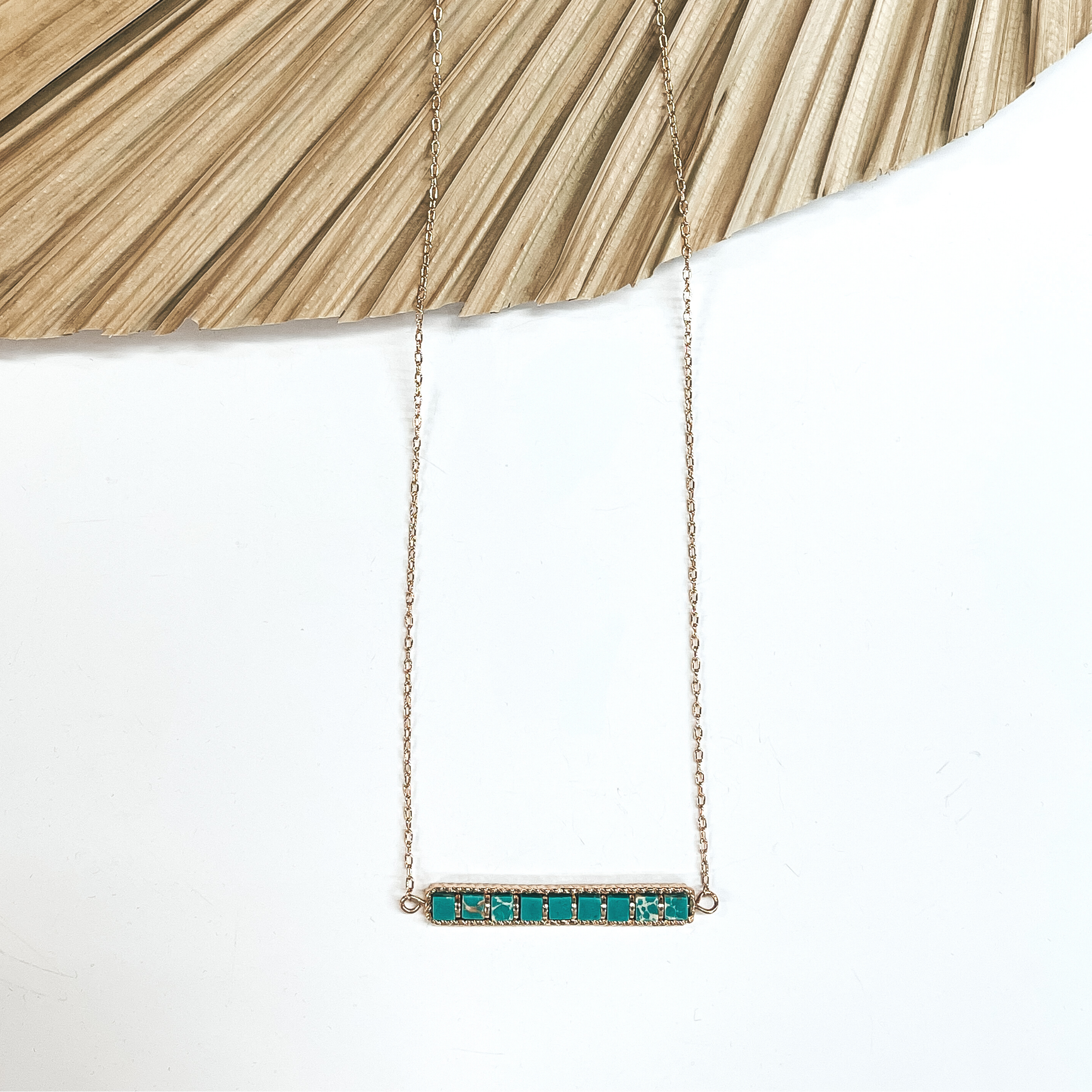This is a gold necklace with a horizontal bar  pendant with semi-precious stones in tuquoise  in a gold setting. This necklace is taken laying on a dried up palm  leaf and white background.